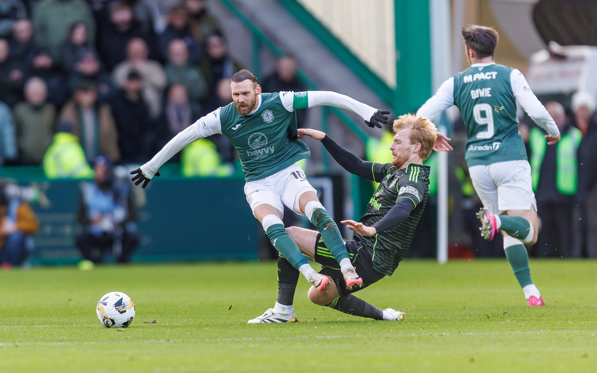 Two soccer players competing for the ball, one in a green and white uniform leaping, the other in black with green accents sliding on the grass. A third player in a green and white uniform runs in the background, and spectators watch from the stands.