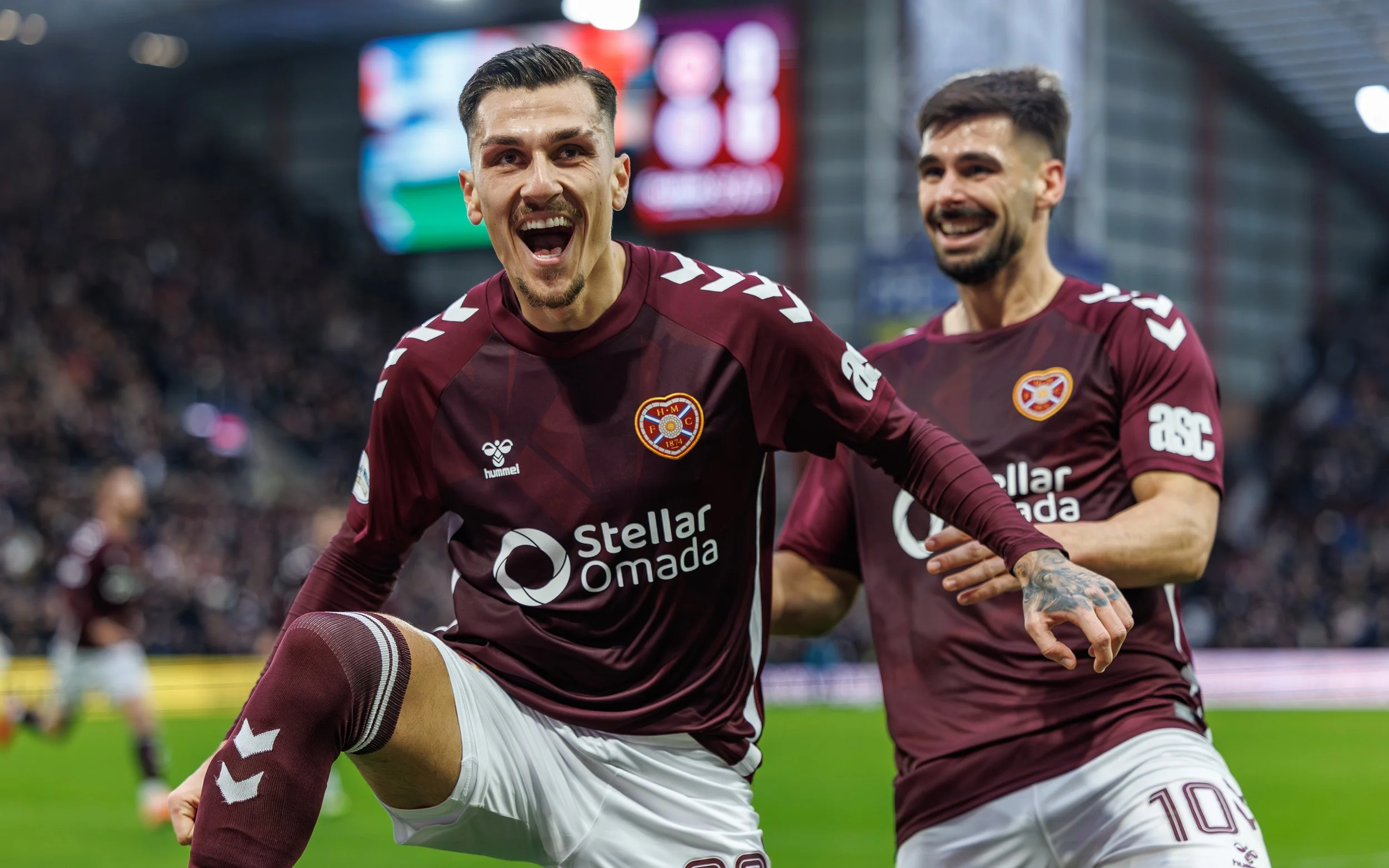 Two soccer players in maroon jerseys celebrating on the field, with one player smiling and having his arm around the other.