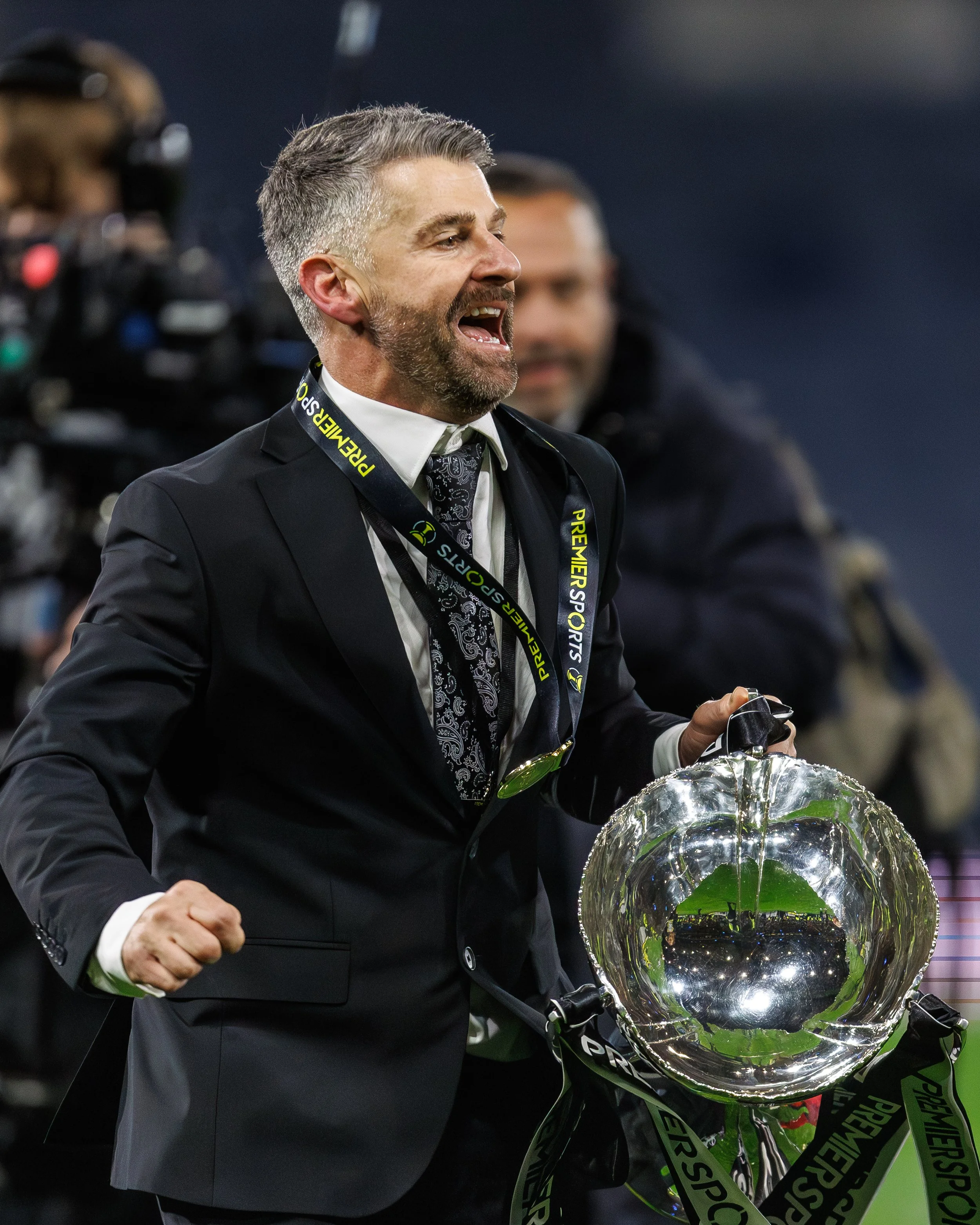 A man in a suit holding a shiny silver trophy, celebrating victory at a sporting event.