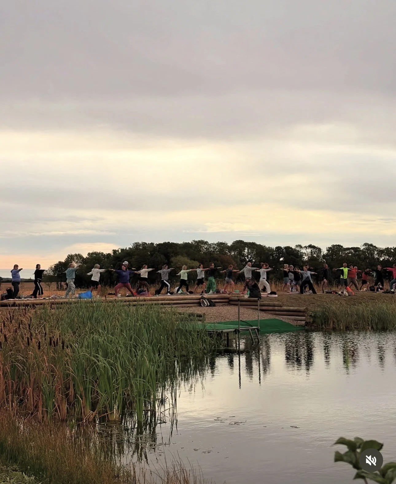 People enjoying a day at a lakeside on a partly cloudy day. Some are swimming, others are preparing for or packing up after swimming. There are backpacks and clothes on the gravel path.