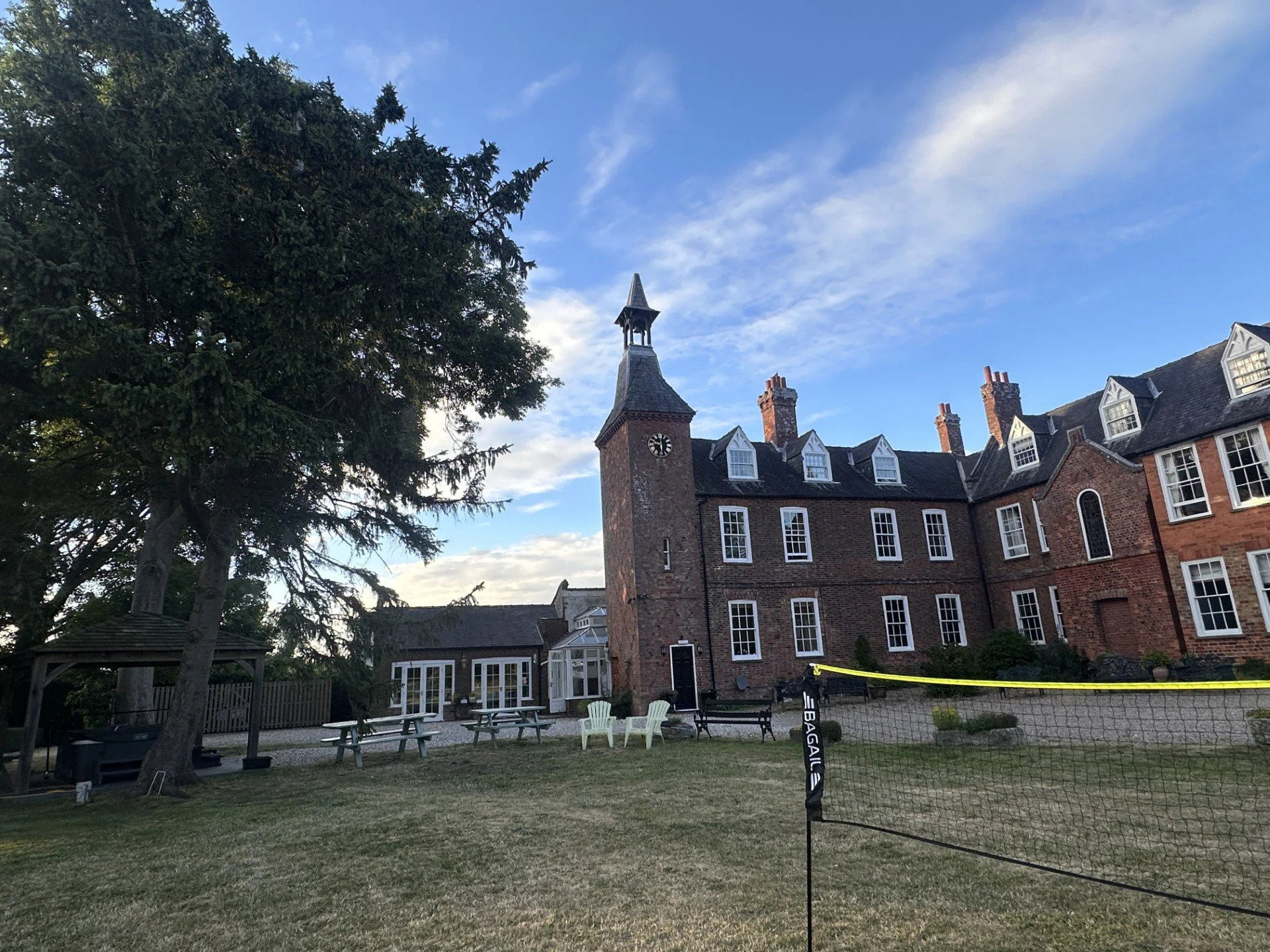 A large, historic brick building with multiple chimneys and dormer windows, next to a tall pine tree under a blue sky with some clouds. There is a grassy area with picnic tables, a volleyball net, and a small wooden gazebo.