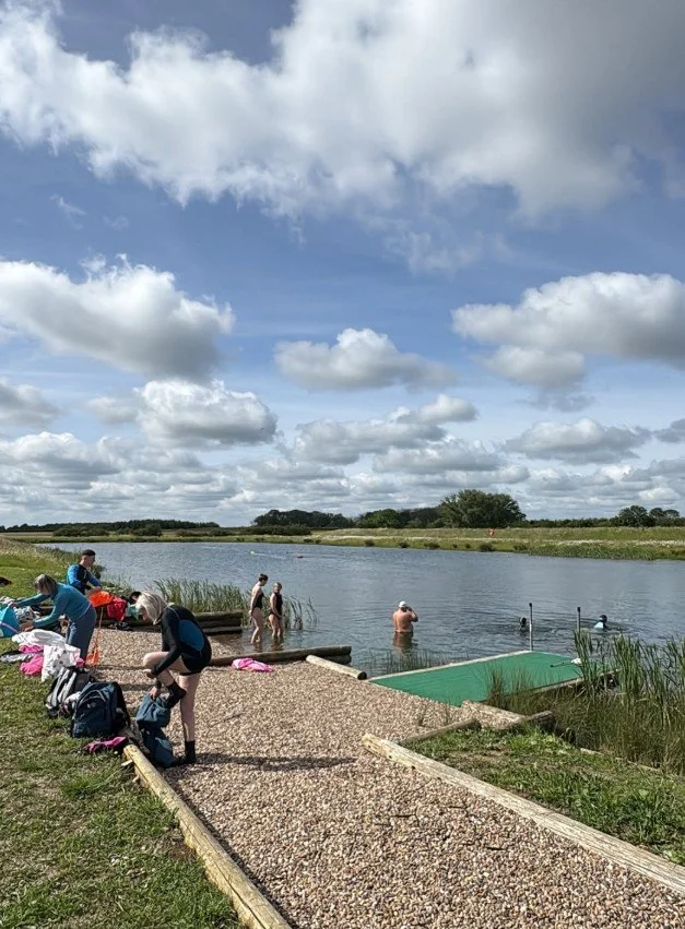 People enjoying a day at a lakeside on a partly cloudy day. Some are swimming, others are preparing for or packing up after swimming. There are backpacks and clothes on the gravel path.