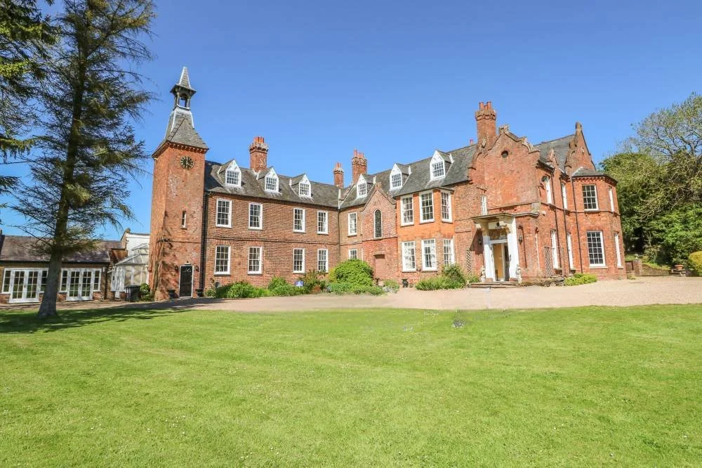 A large, historic brick mansion with a tall tower on the left, multiple windows, and a well-maintained lawn in front, under a clear blue sky.