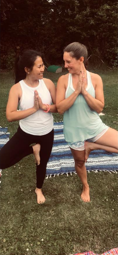 Amanda practicing yoga outdoors with a client on a grassy field with trees in the background, standing on a striped blanket, smiling and facing each other with palms together in a prayer pose.
