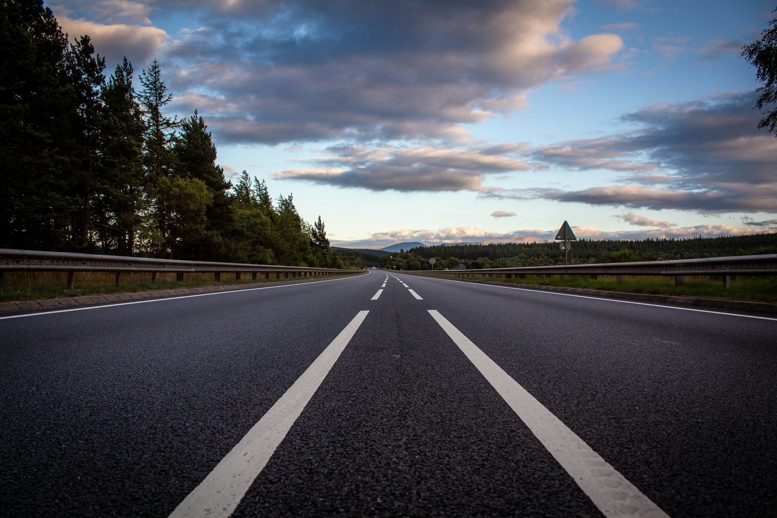 Empty two-lane road with guardrails on both sides, trees along the edges, and a cloudy sky during sunset.