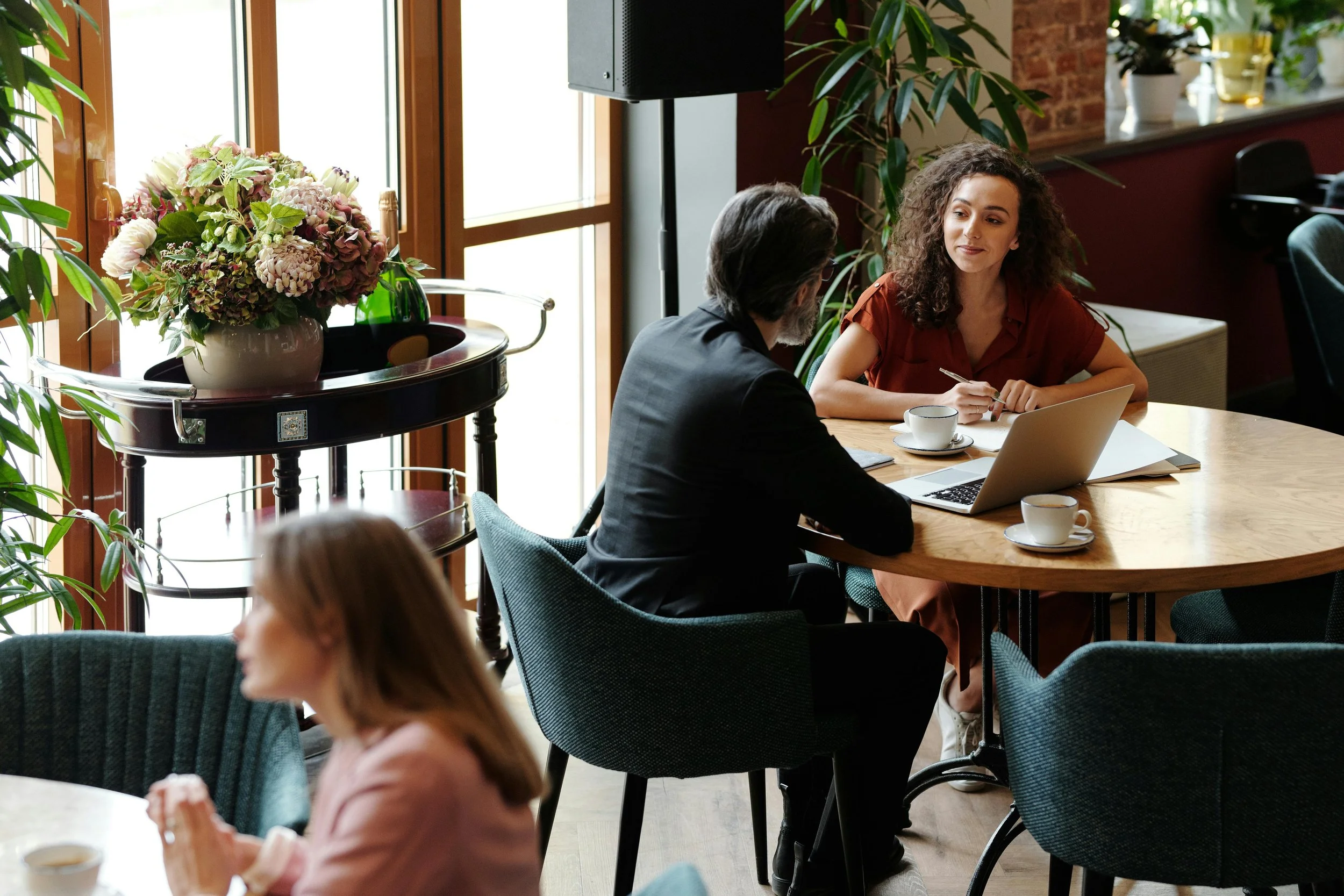 Two people are sitting at a round wooden table in a cozy cafe or meeting space, engaged in a conversation with laptops, notebooks, and coffee cups. In the foreground, a woman with red hair is slightly out of focus, while in the background, a man with dark hair and a woman with curly hair are actively talking.