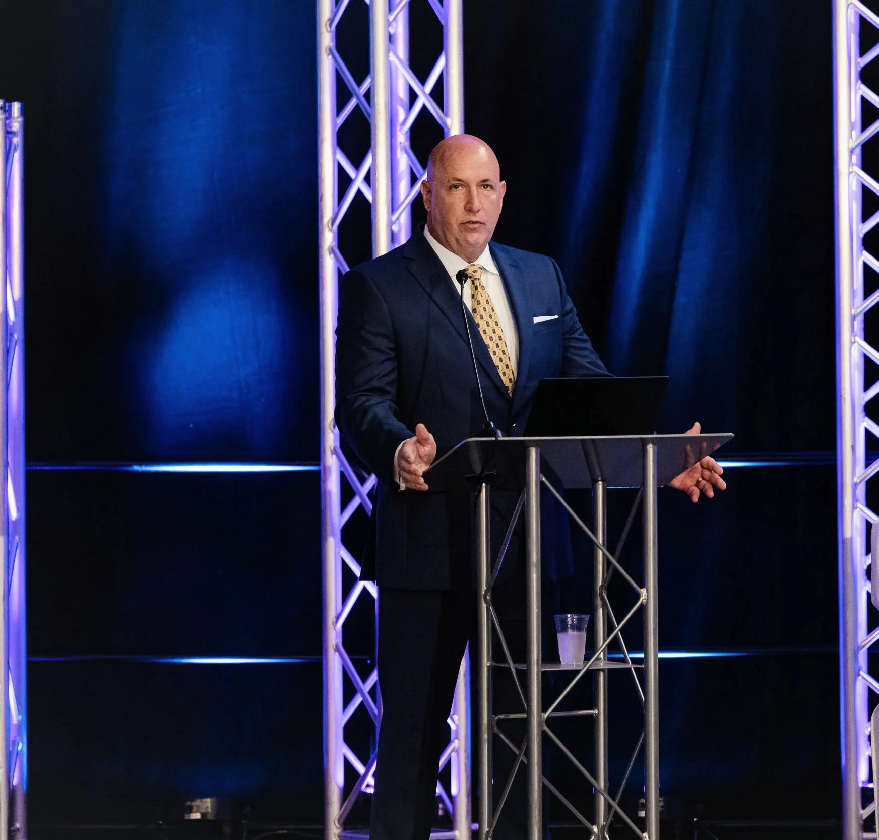 A man in a dark suit and patterned tie stands at a podium with a laptop, speaking at an event with blue lighting and stage truss in the background.