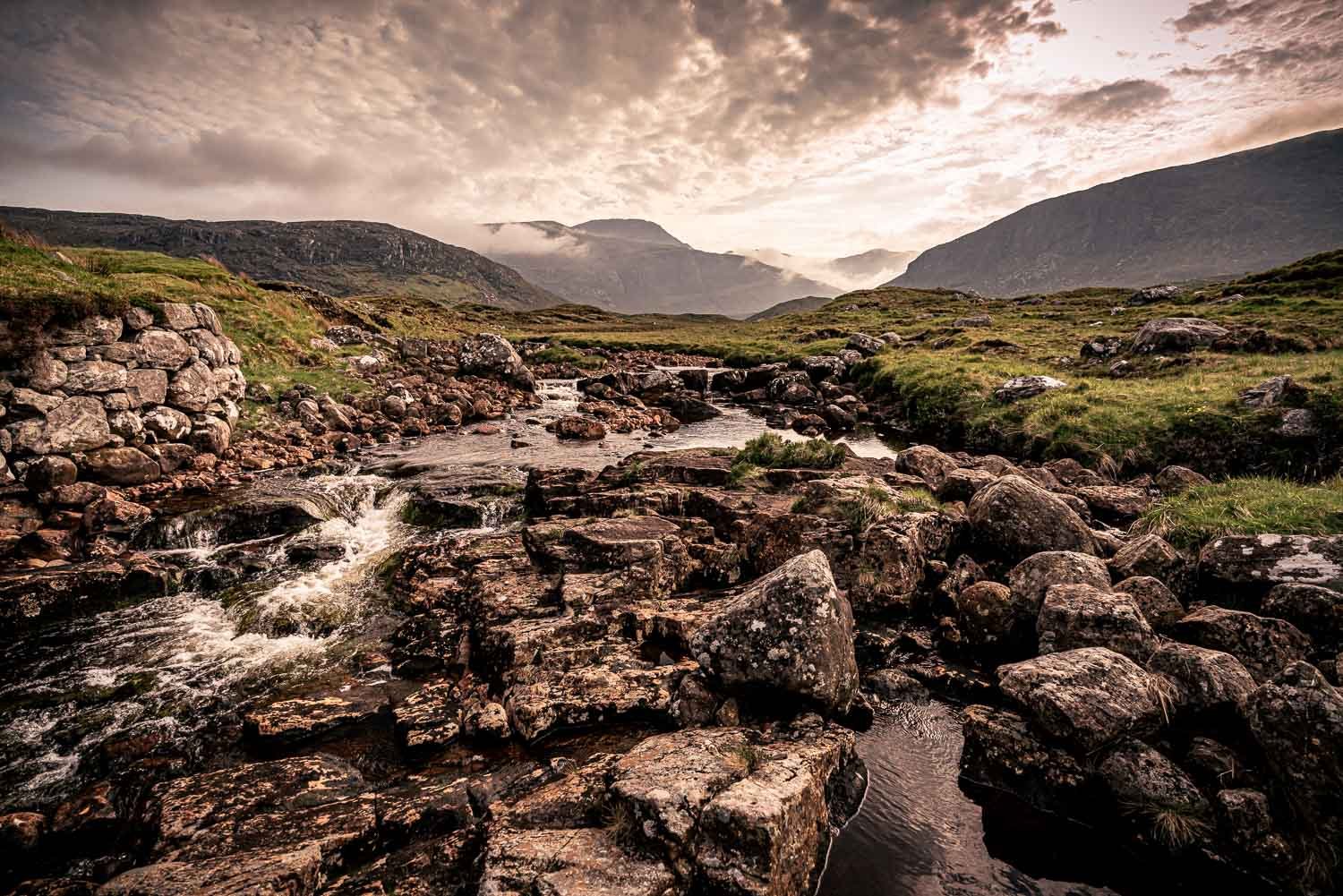 A scenic mountain landscape with a rocky stream flowing through grassy moorland terrain under a dusky and cloudy sky.