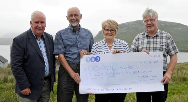 Four people standing outdoors on a grassy hill holding a large check made out to Crossroads Harris for fifteen thousand pounds, with a loch and mountains in the background.