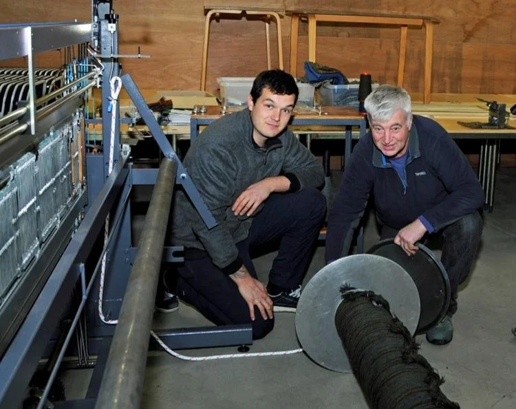 Two men in a weaving shed with a loom, working on a large beam of yarn ready for weaving.