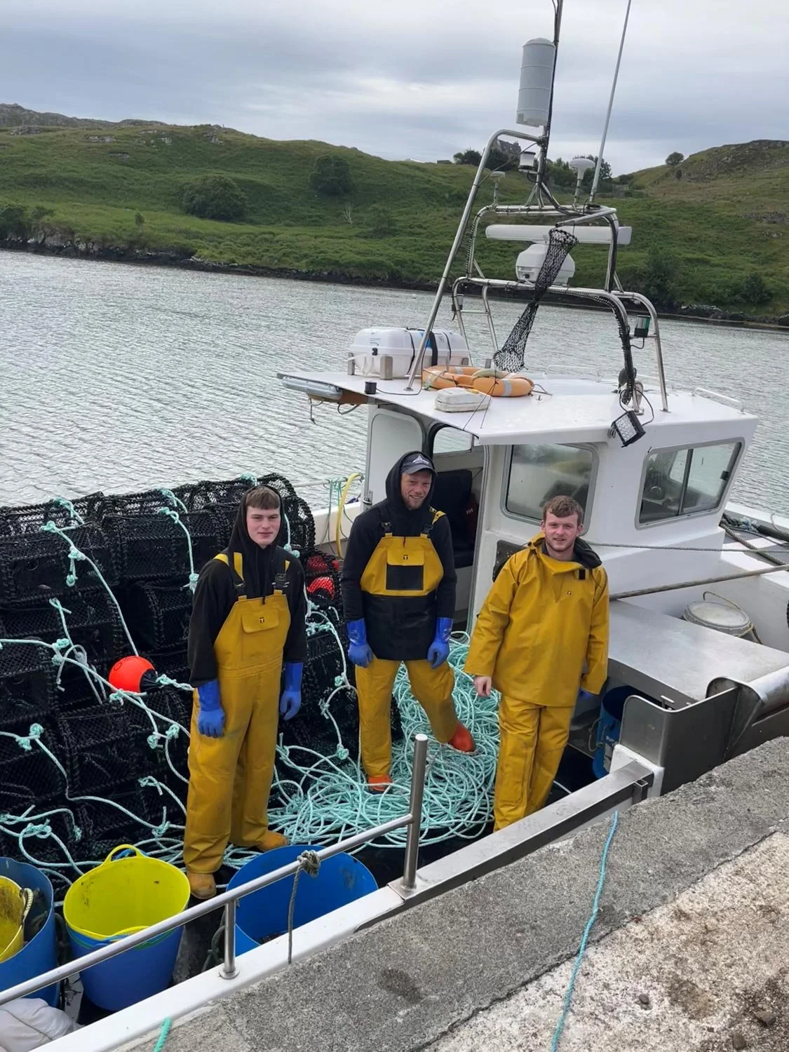 Three fishermen in yellow waterproof gear on a boat with crab traps and orange buoys, near a rocky shoreline with green moorland in the background.