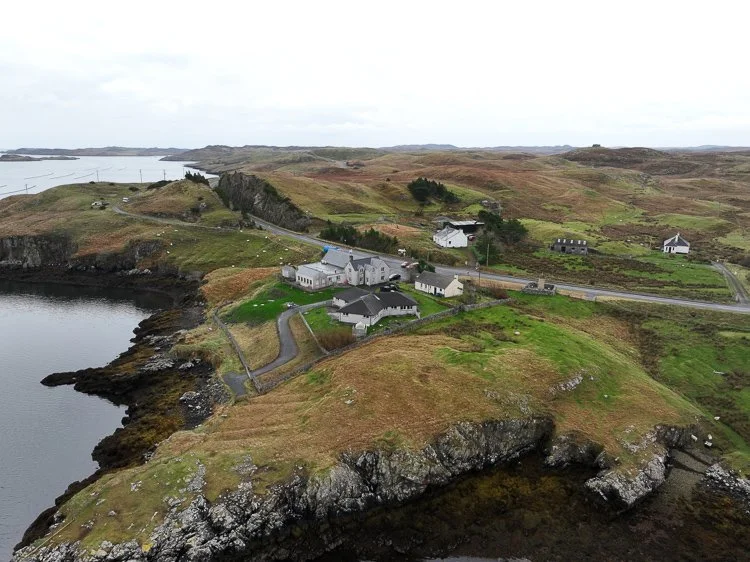 A coastal village with a few houses and community buildings, surrounded by cliffs and green crofts, overlooking the loch under a cloudy sky.