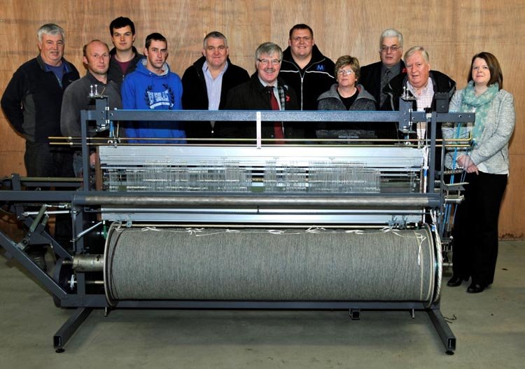Group of eleven people standing behind large industrial double width loom in a weaving shed.