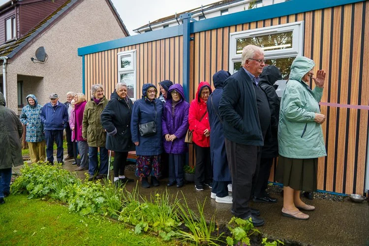 People standing in line outside a building with a wooden panel exterior on a rainy day, some wearing raincoats and holding umbrellas.