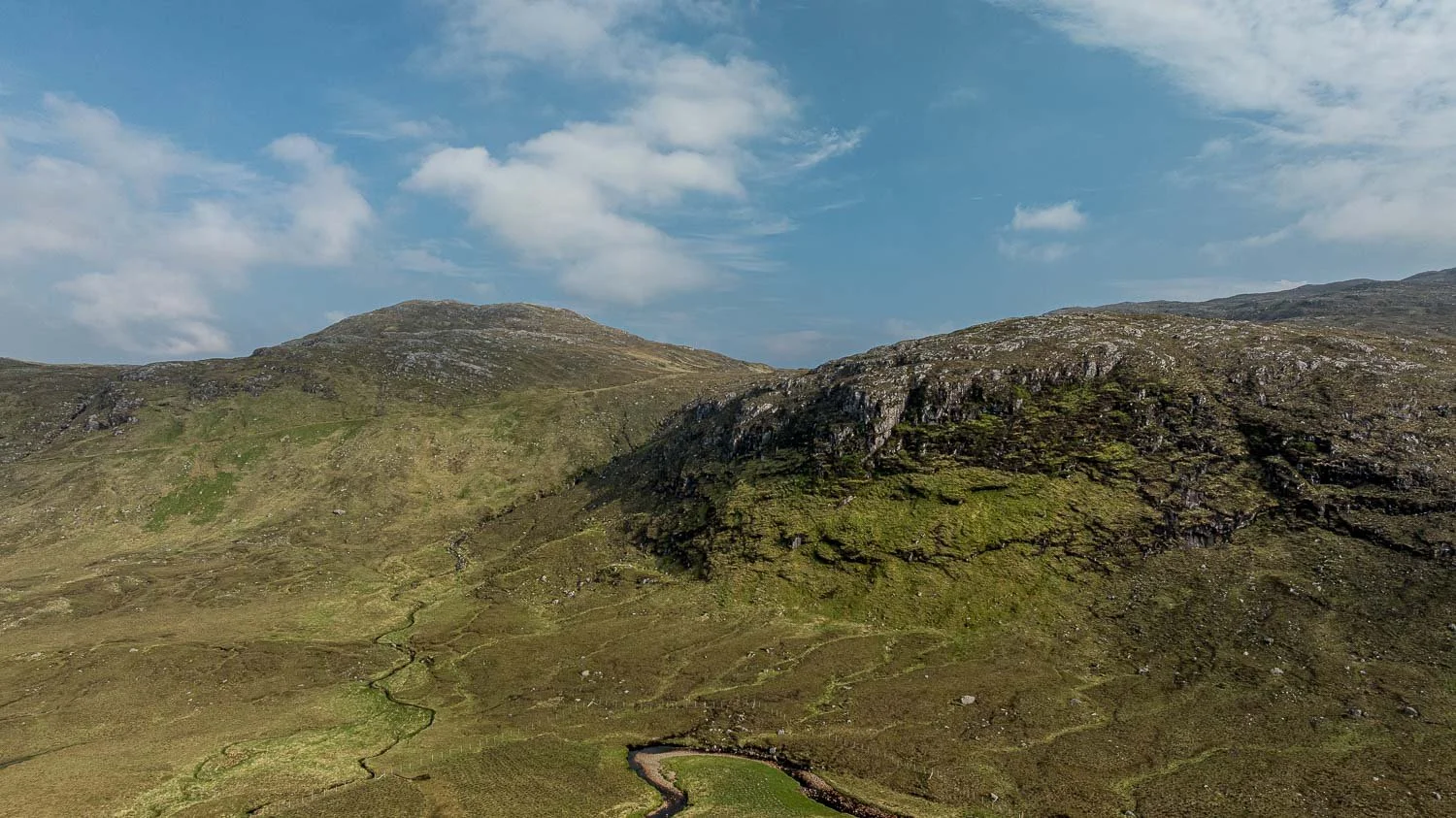 A panoramic view of rolling green hills with rocky outcroppings under a partly cloudy sky.