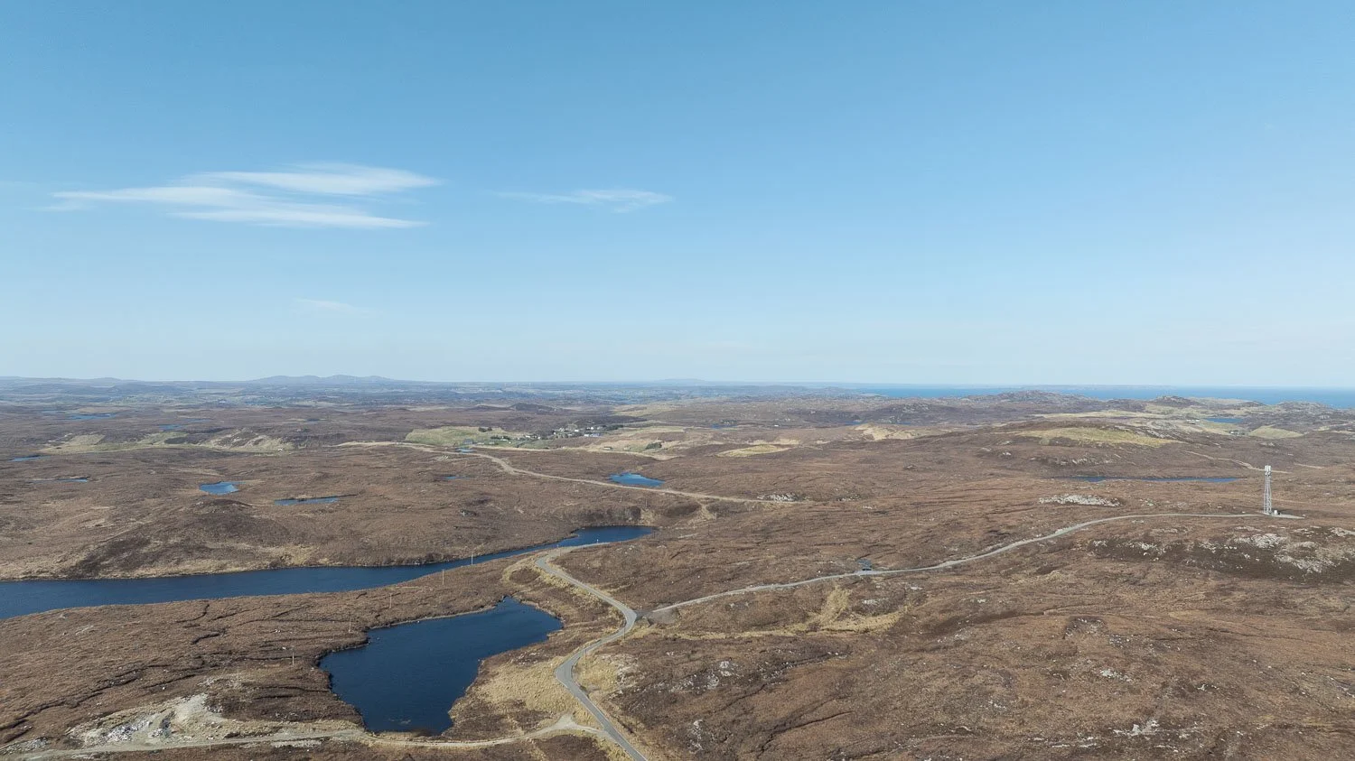 Aerial view of a vast, open landscape with brown moor, several lochs, and a winding road under a clear blue sky.