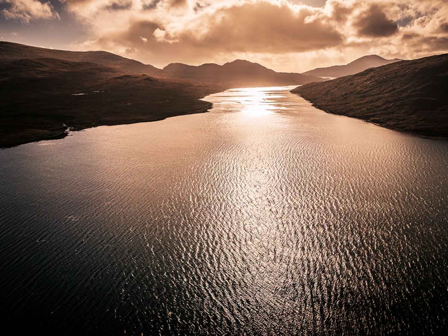 A serene body of water, a loch in the Outer Hebrides, reflecting the light of the setting or rising sun, surrounded by mountains under a partly cloudy sky.