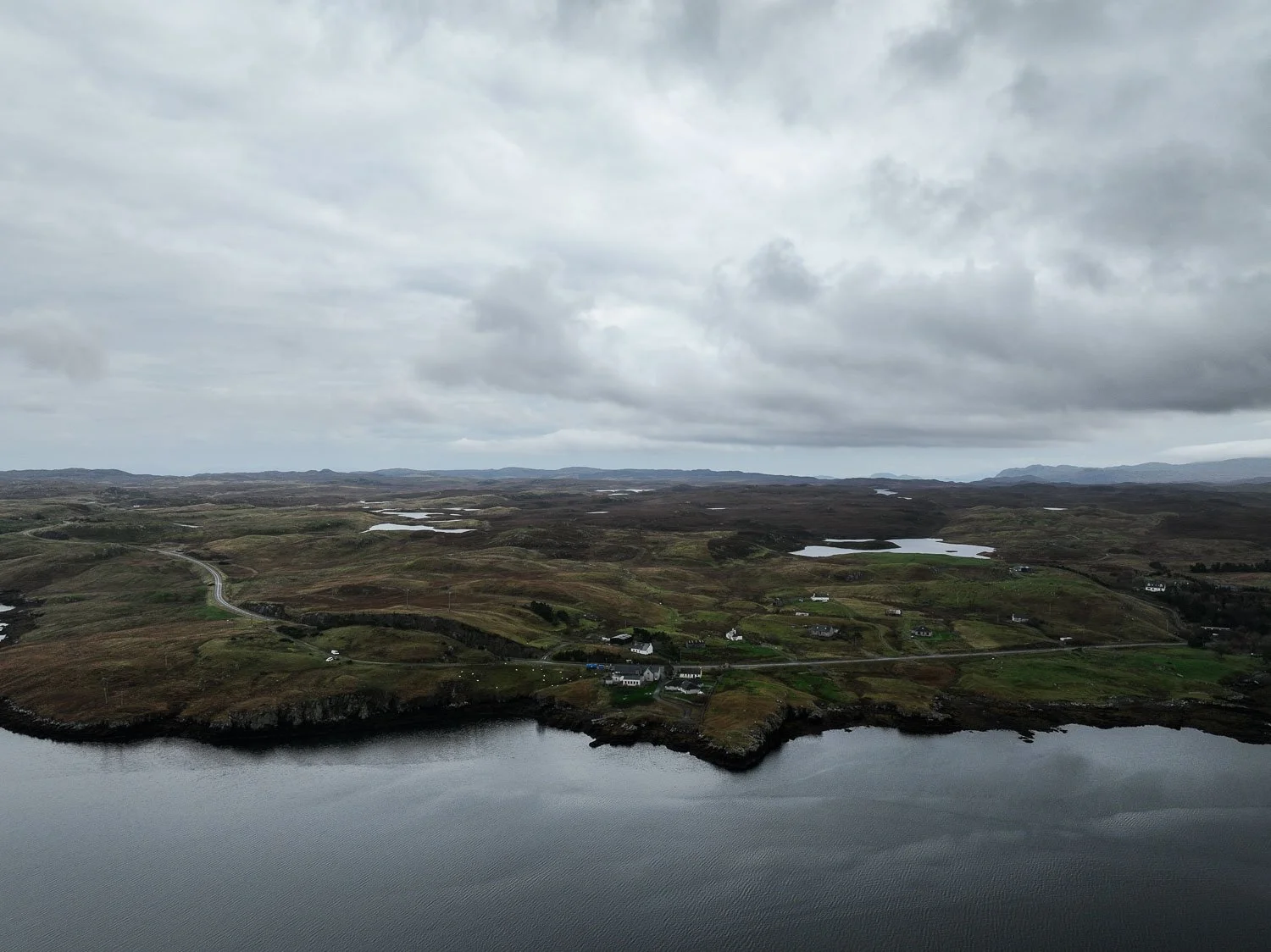 Aerial view of a rugged landscape with a body of water in the foreground, expansive grassy hills, villages, and small lochs under a cloudy sky.