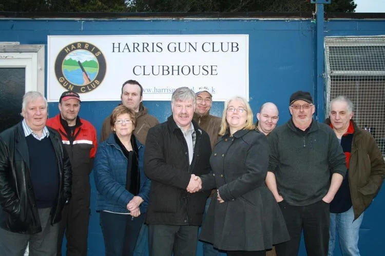 Group of nine people standing in front of a blue wall with a sign that reads "Harris Gun Club Clubhouse." Two people in the front are shaking hands and smiling. The group is casually dressed.