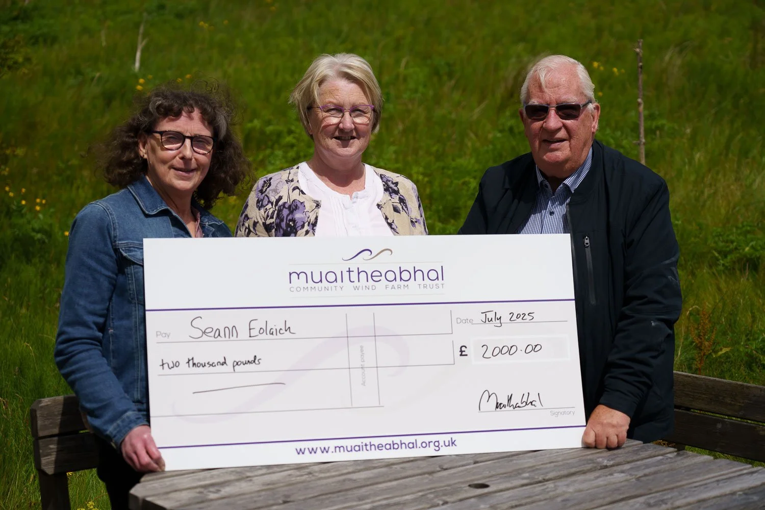 Three people standing outdoors holding a large cheque made out to Seann Eolaich for two thousand pounds from Muaitheabhal Community Trust, in July 2025.