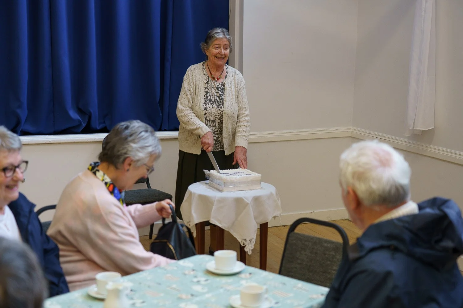 A woman with grey hair is cutting a cake at a gathering, surrounded by seated people at a table.