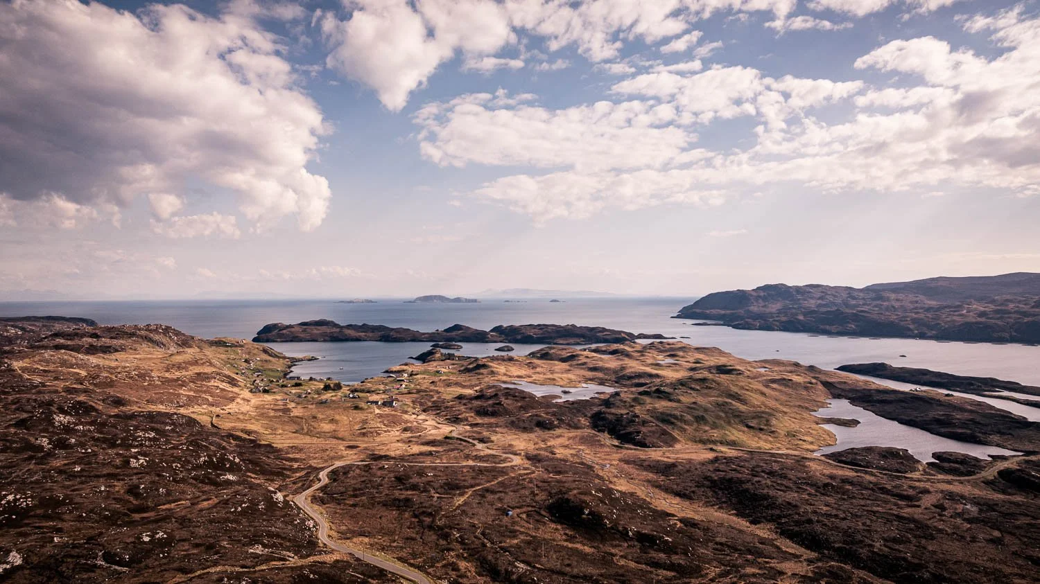 Aerial view of a coastal landscape with hills, loch entrance, and small houses, under a partly cloudy sky.