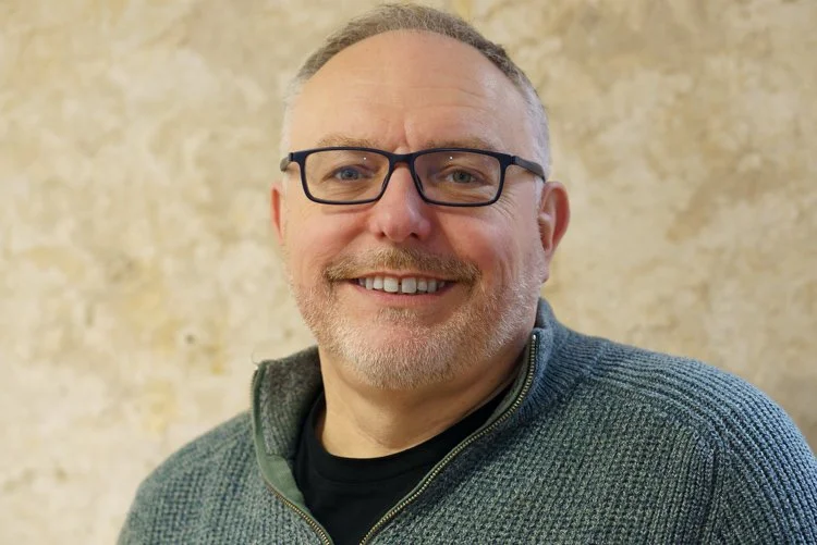 A man with glasses, smiling, wearing a grey sweater over a black shirt, standing in front of a beige textured wall.