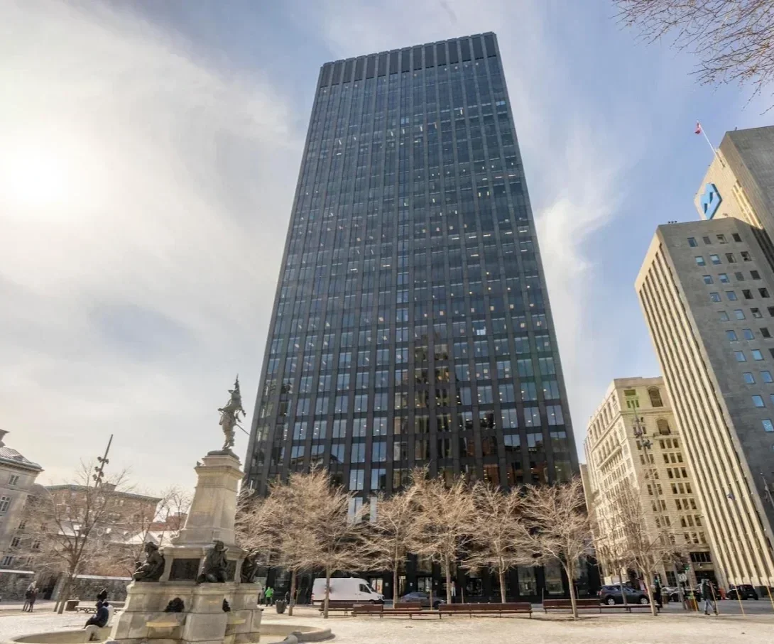 Tall black office building with multiple windows, surrounded by shorter buildings and leafless trees in a city square with a monument at the front.