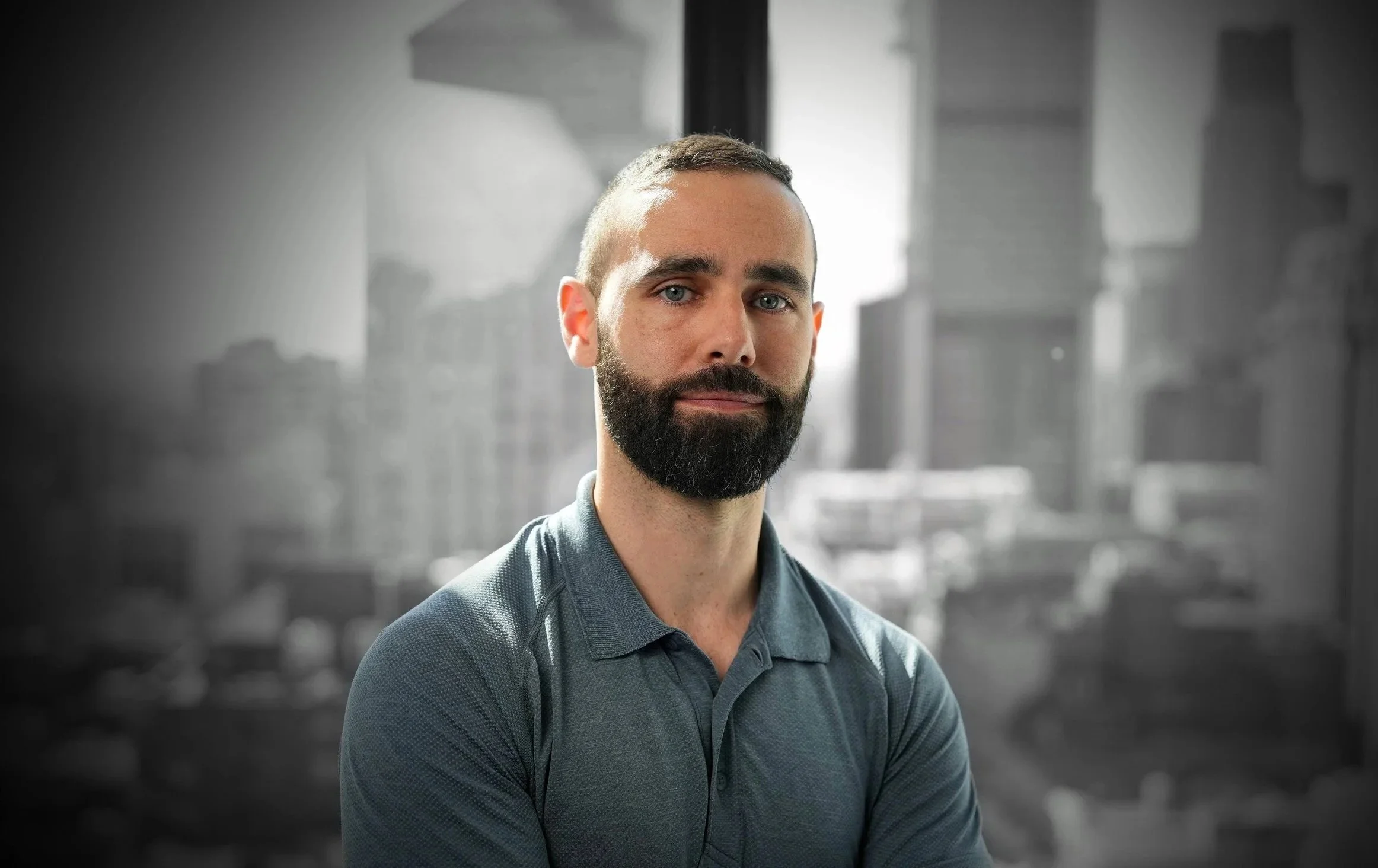 Portrait of a man with a beard and short hair, wearing a collared shirt, standing in front of a blurred cityscape background.