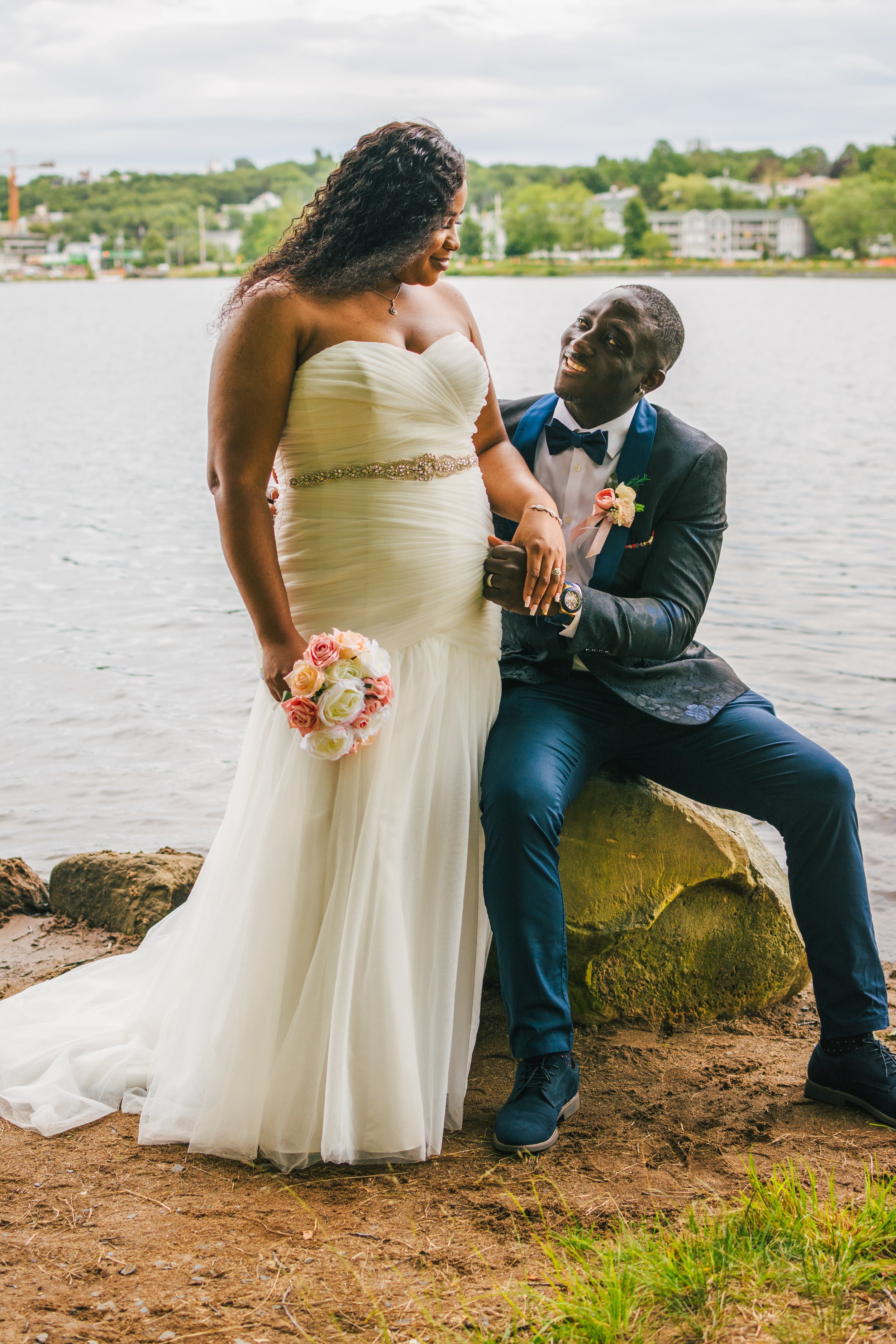 A bride and groom by a lake, with the groom kneeling on a rock, smiling at her as she holds a bouquet of pink and white roses.
