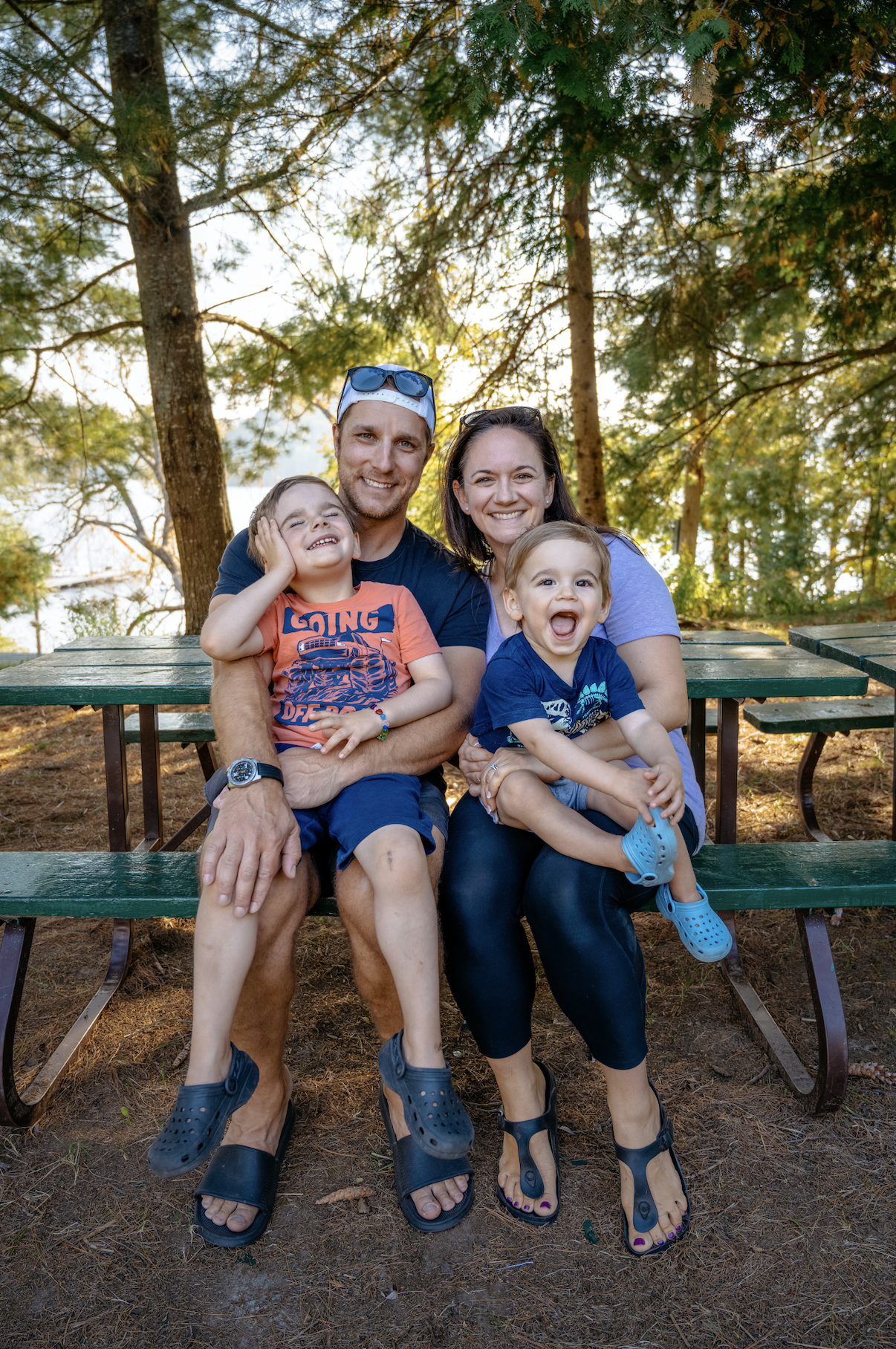 A happy family of four sitting on a park bench in a wooded area with tall trees, smiling and enjoying outdoor time.