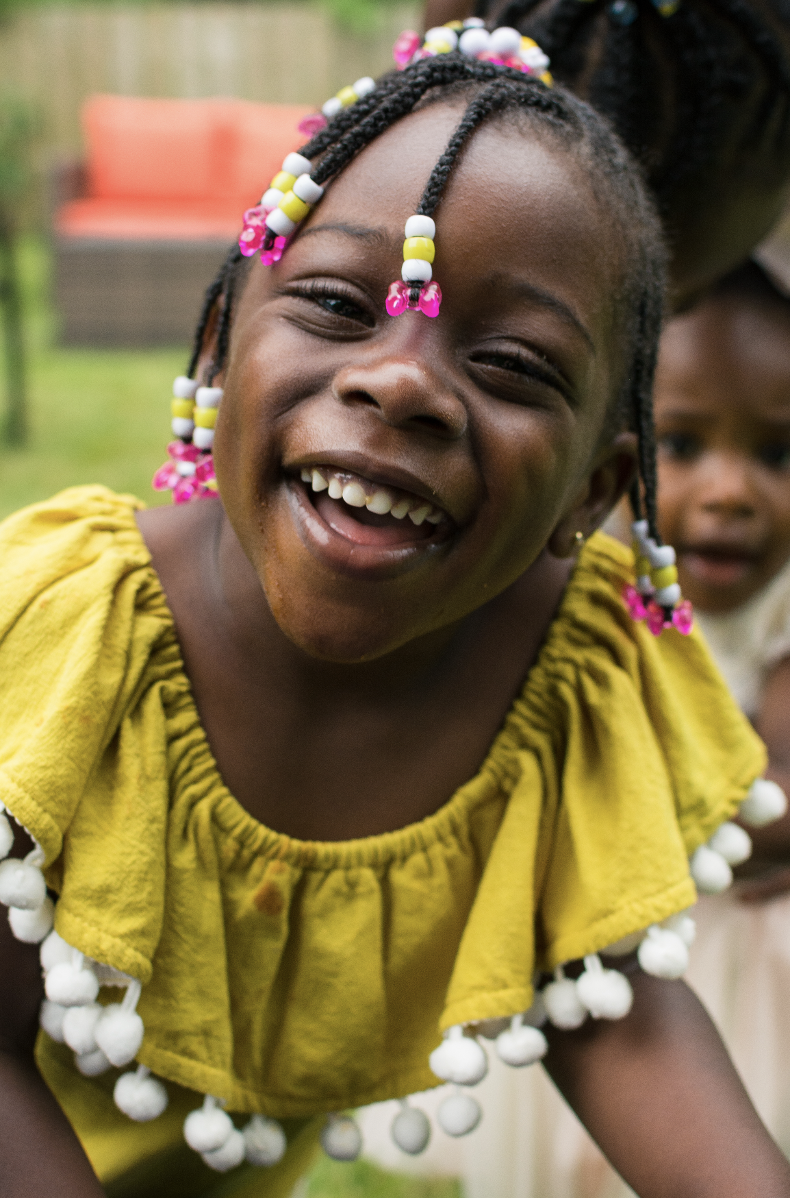 A young girl smiling and laughing, wearing a yellow dress with white pom-poms, with braided hair adorned with pink, yellow, and white beads, outdoors.