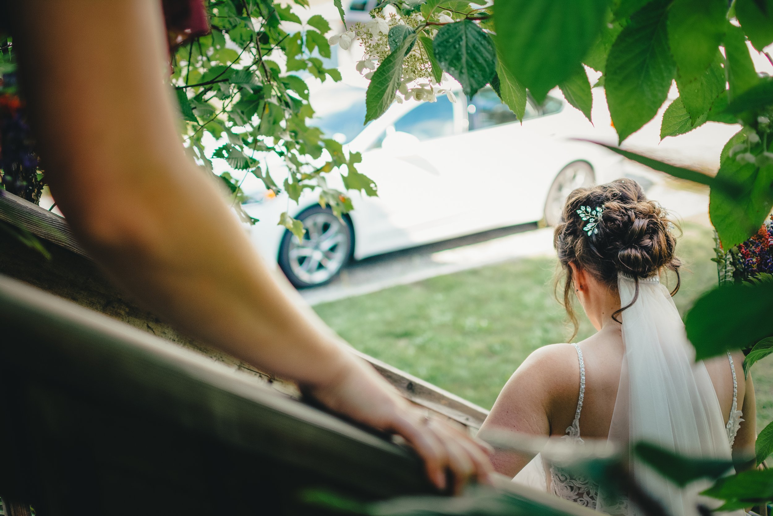 Bride with braided hair, wearing a white wedding dress and veil, viewed from behind, surrounded by greenery, with a white car in the background.