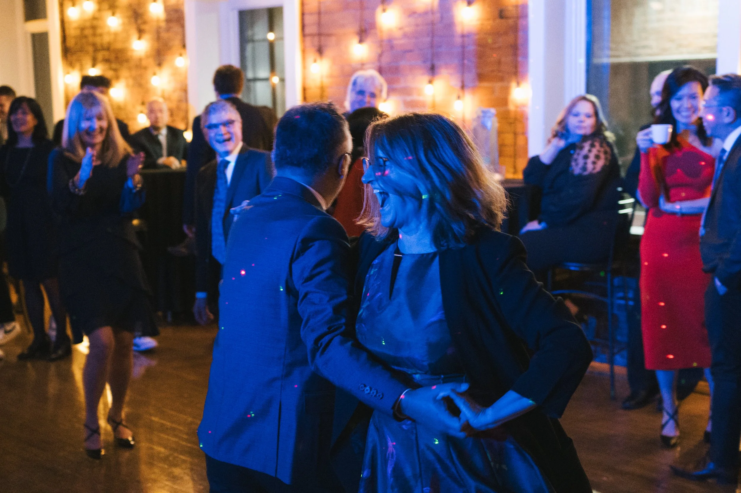 People dancing and socializing at an indoor party or event, with warm lighting and brick walls in the background.