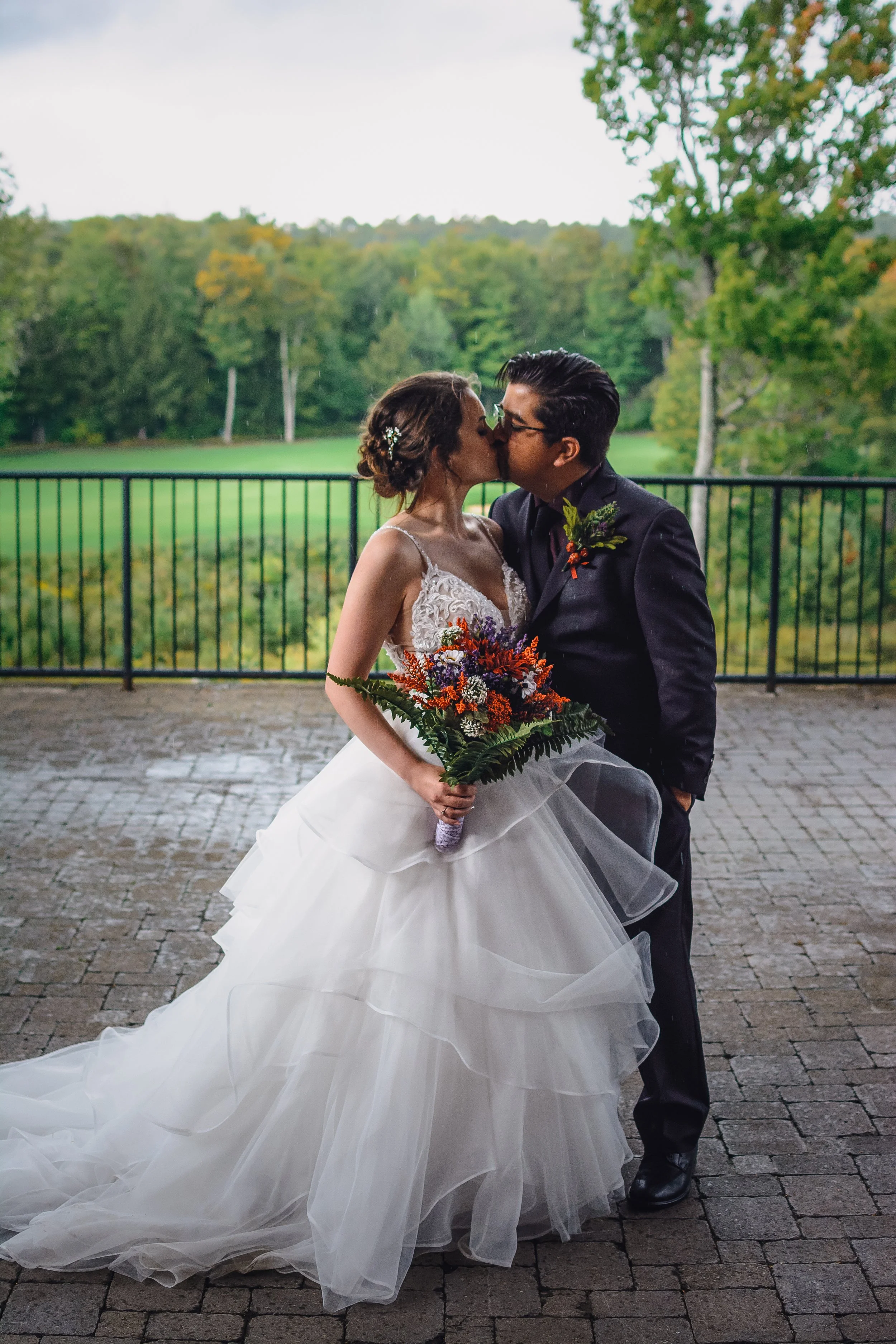 A bride and groom share a kiss on their wedding day outdoors with a green landscape and trees in the background.
