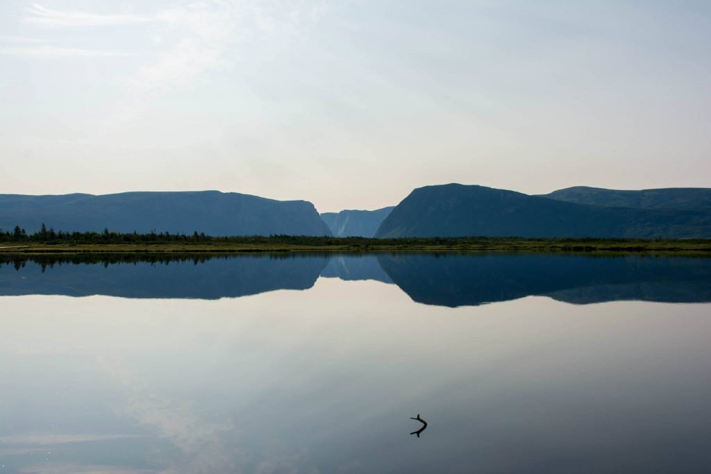 A calm lake reflecting mountain ranges and a partly cloudy sky, with a kayak paddle floating in the water.