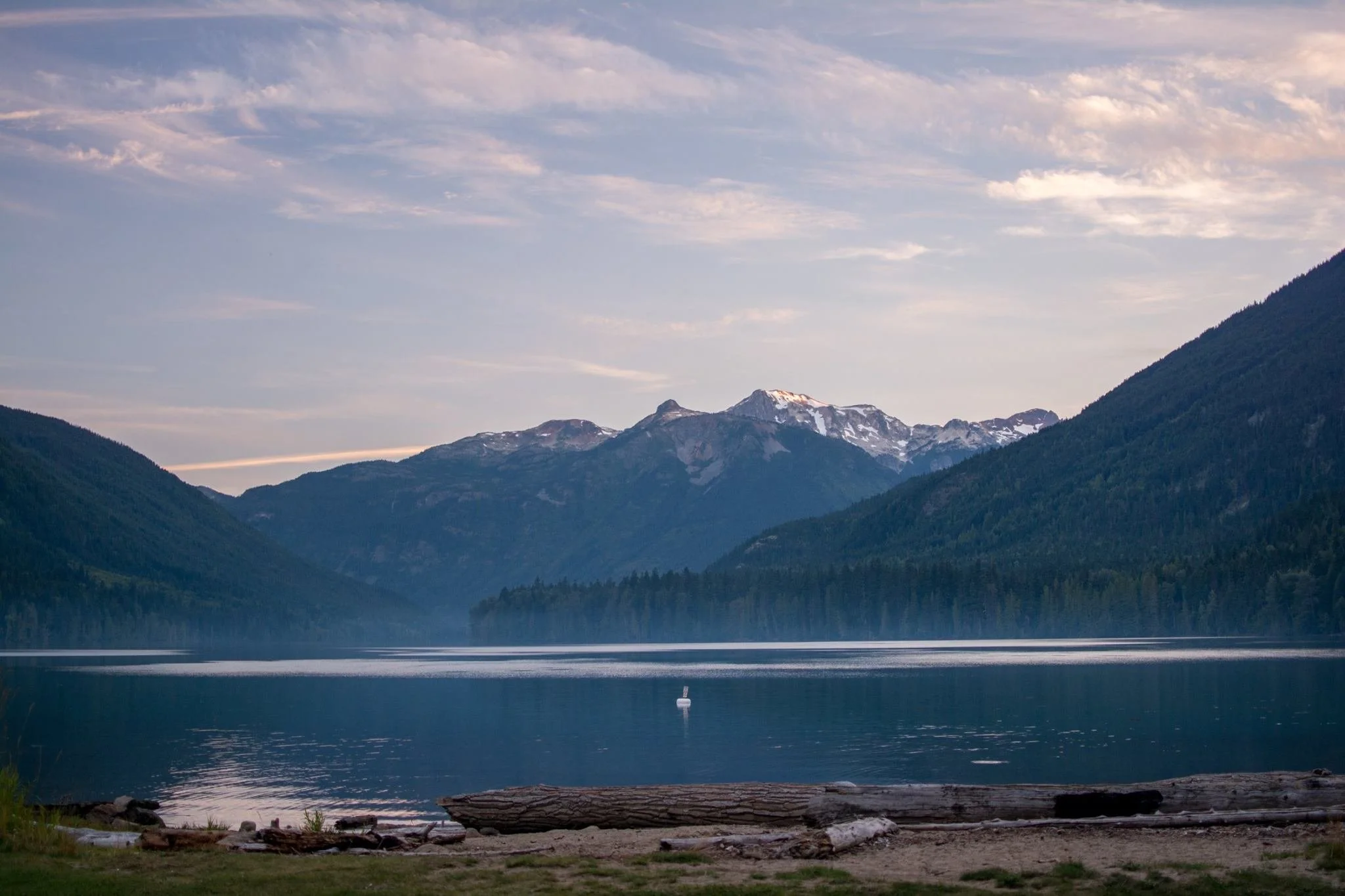 Scenic view of a large mountain lake surrounded by forested mountains, with snow-capped peaks in the background, a partly cloudy sky, and a swan swimming in the water.