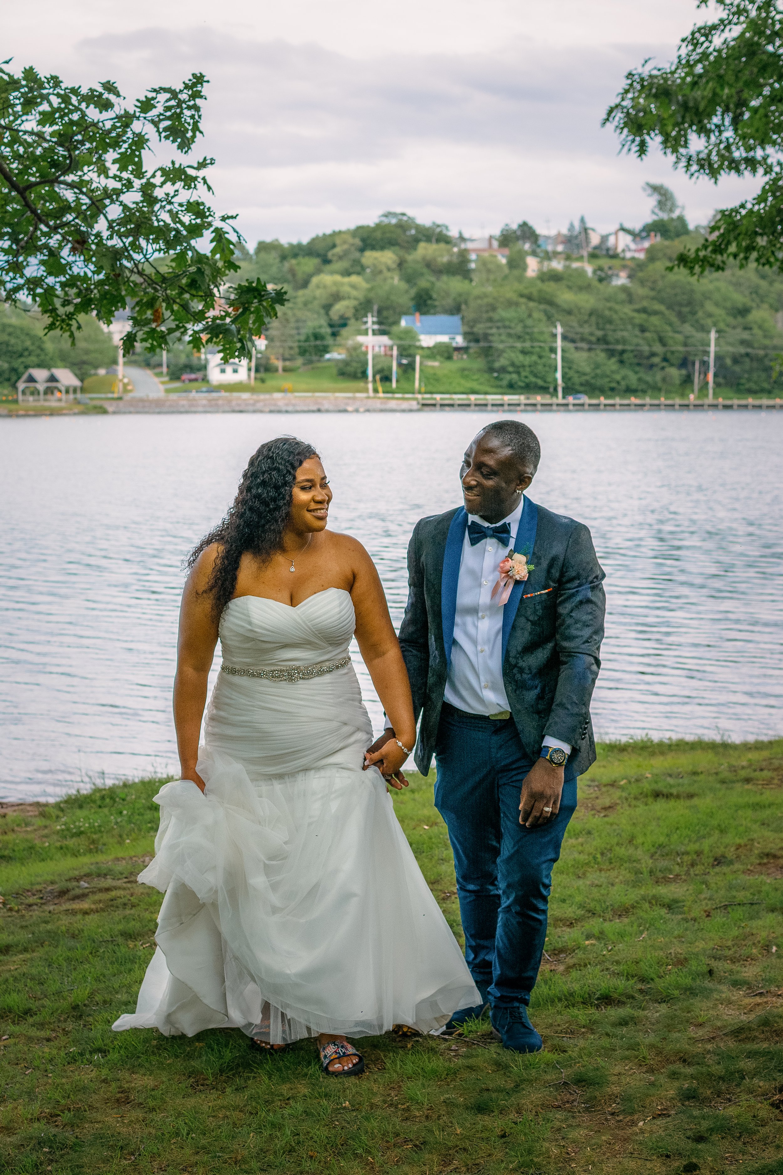 Couple walking by a lake, with the bride in a strapless white wedding dress and the groom in a dark suit with a blue bow tie, holding hands and smiling at each other.