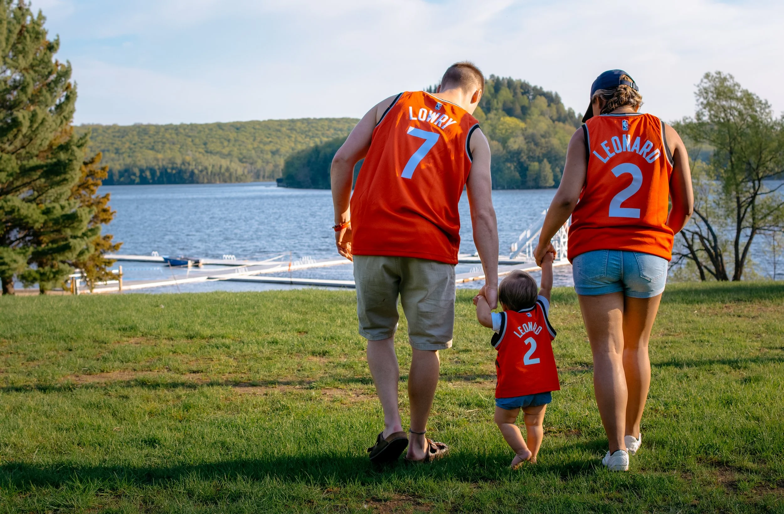 A family of three holding hands and walking on grass near a lake. They are wearing matching red basketball jerseys with the names and numbers Lowry 7, Leonard 2, and Leonard 2 for the child. The background features a lake, trees, and a clear sky.