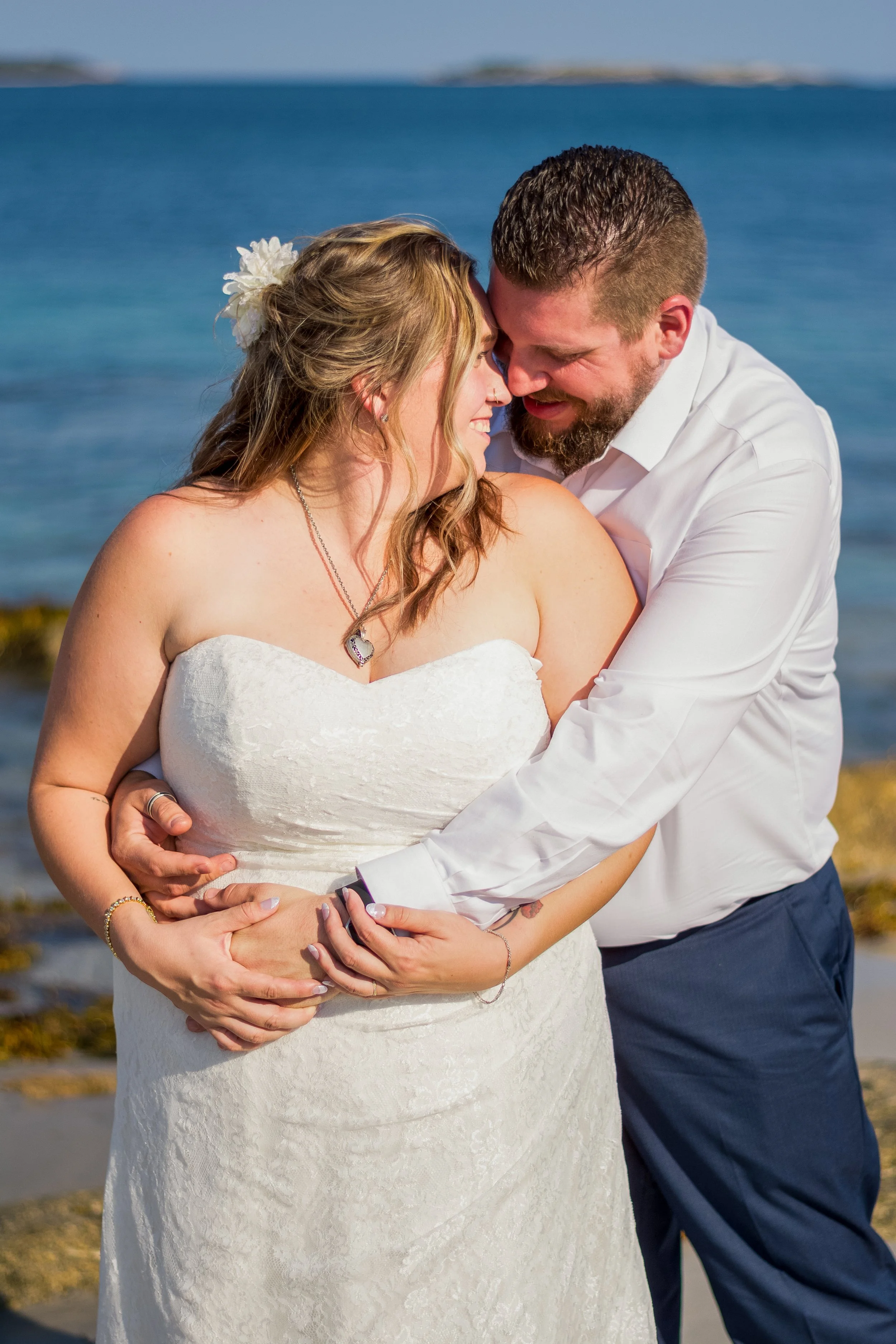 A couple in wedding attire embracing on a beach with the ocean in the background.