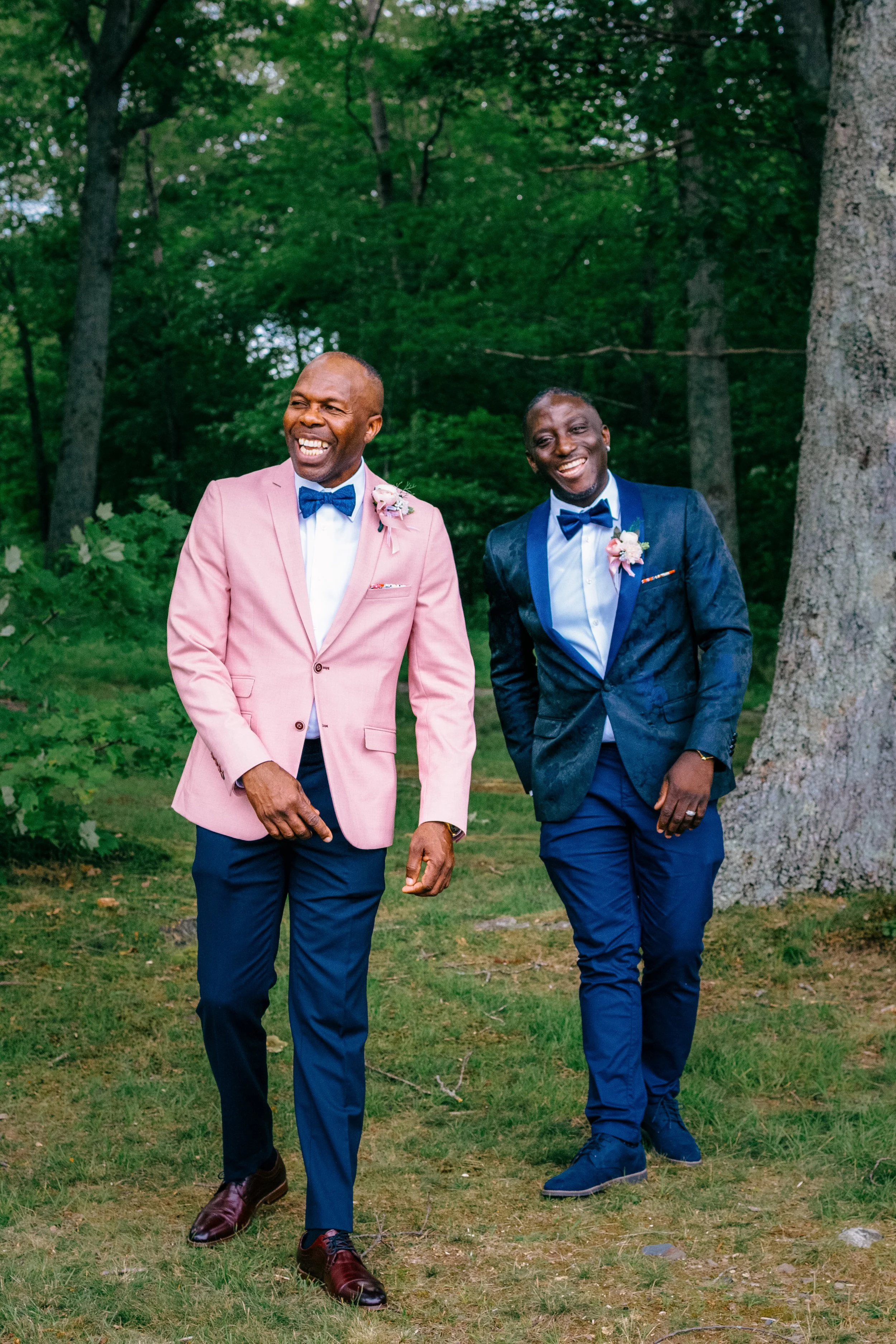 Two men dressed in suits and bow ties, walking outdoors in a green wooded area, smiling and laughing.
