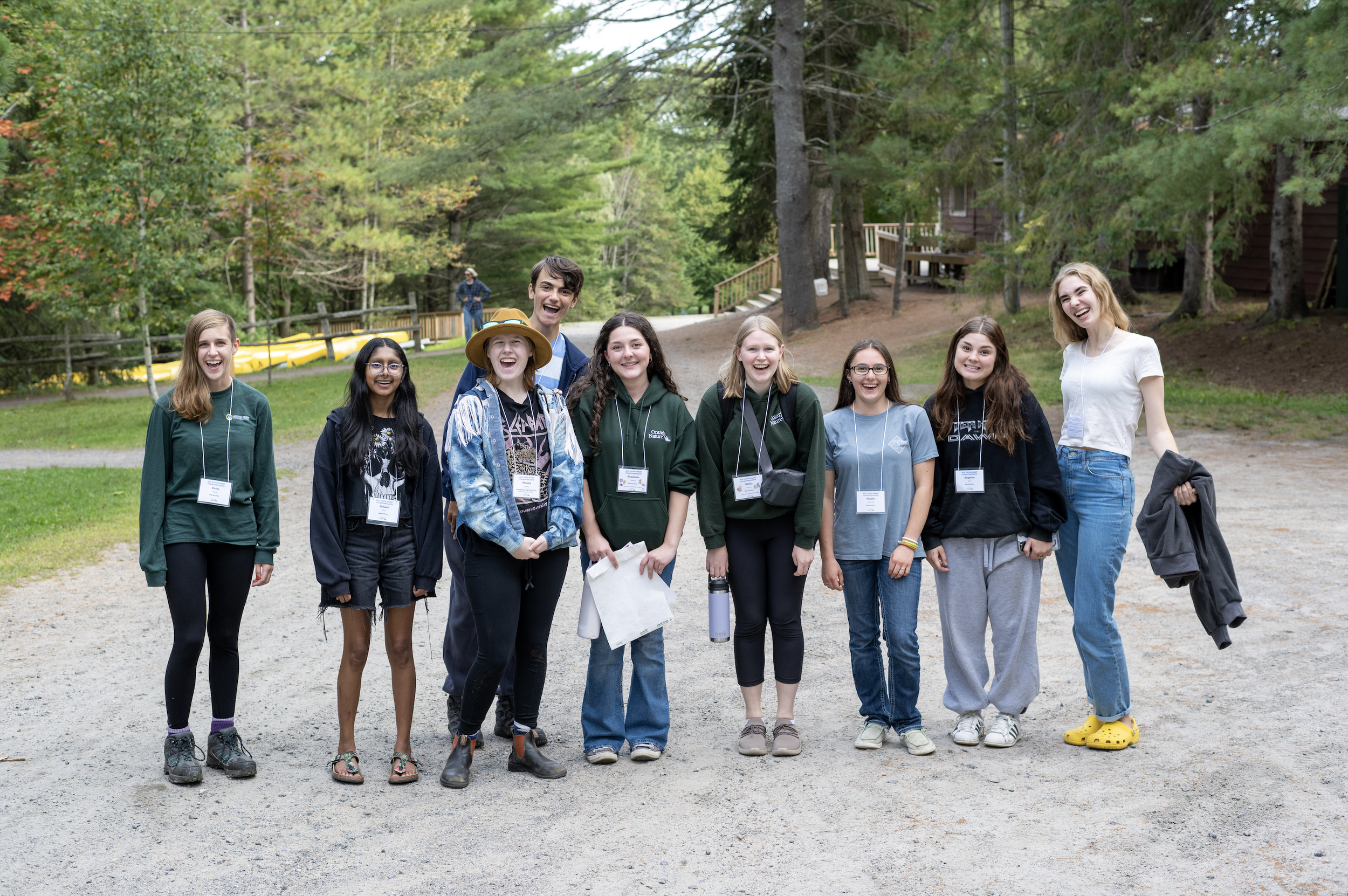 Group of nine teenagers standing outdoors on a gravel path, smiling and laughing, with trees and wooden cabins in the background.