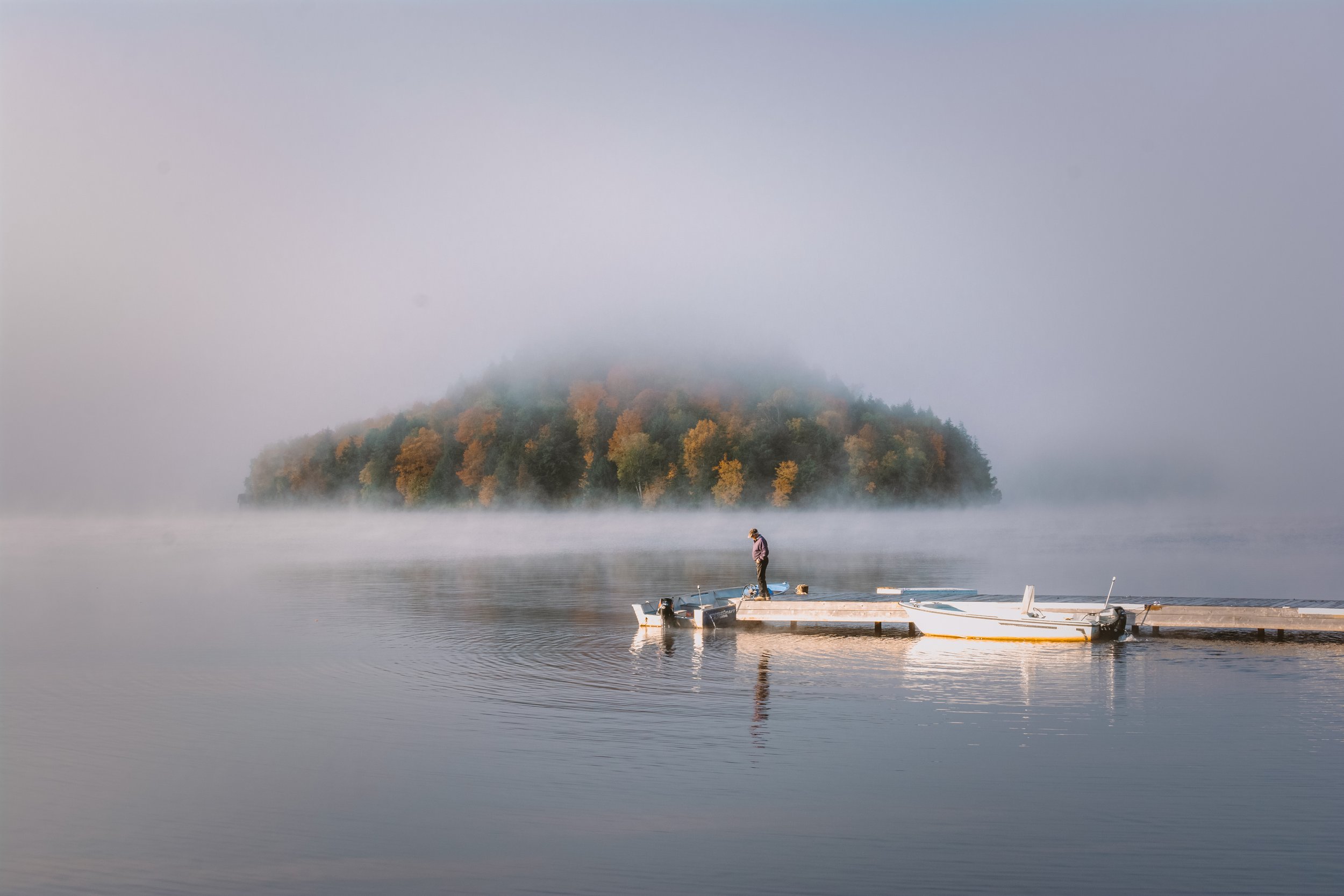 A person standing on a wooden dock by a calm lake with a foggy island in the background.