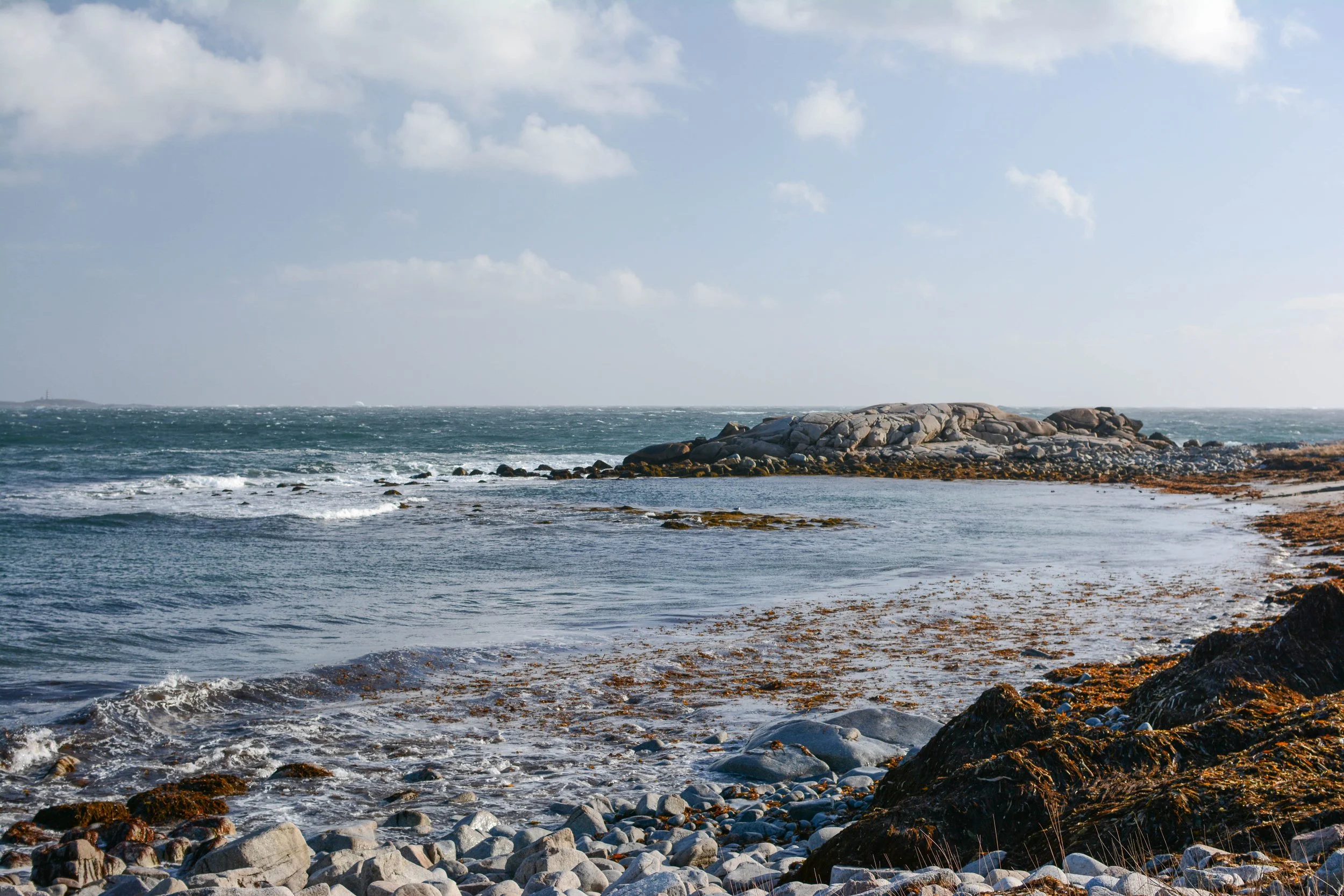 A rocky shoreline with a calm ocean and a large rock formation in the background under a partly cloudy sky.