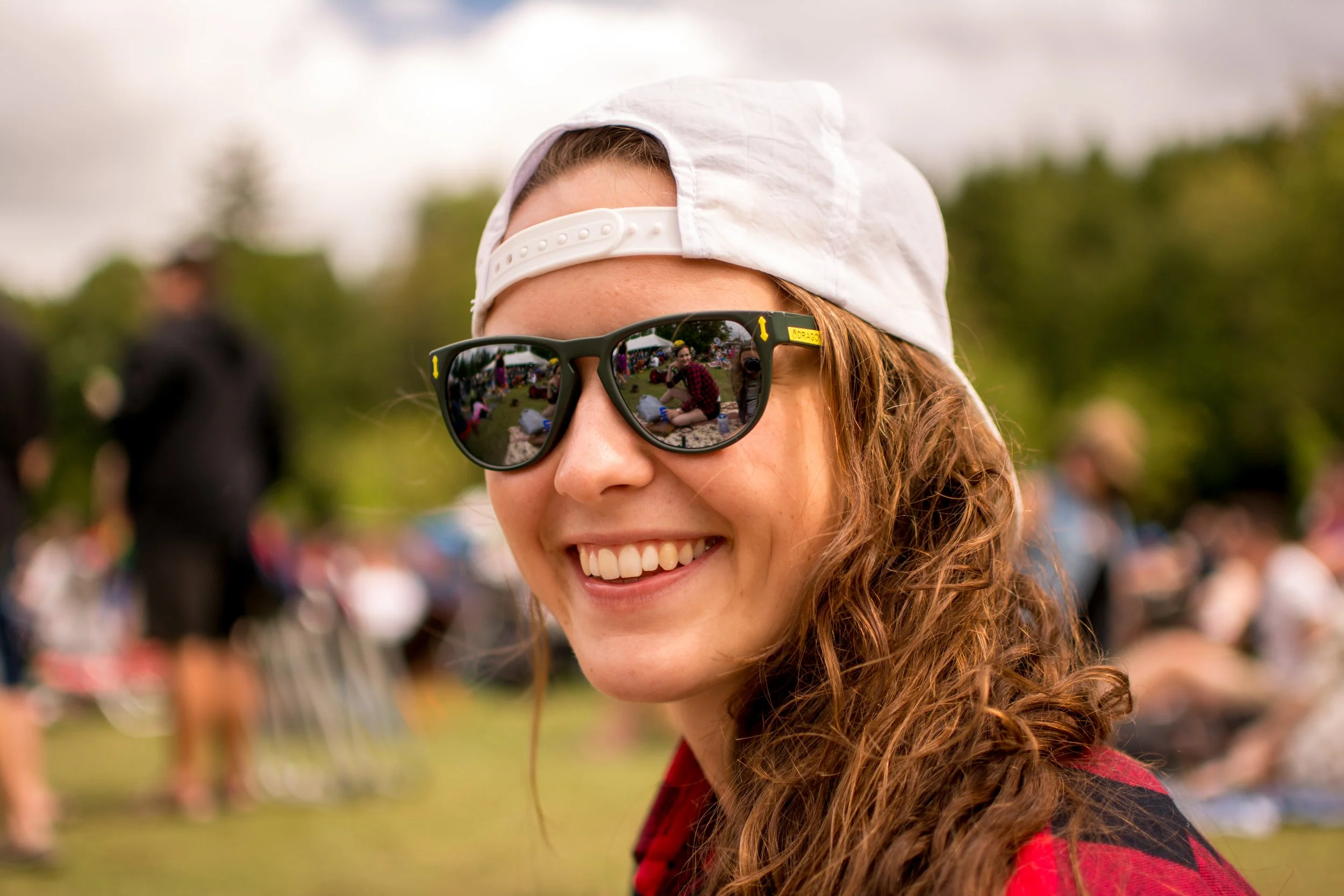 Young woman with curly red hair wearing sunglasses, a white cap backwards, and a red shirt, smiling at an outdoor gathering with people and trees in the background.