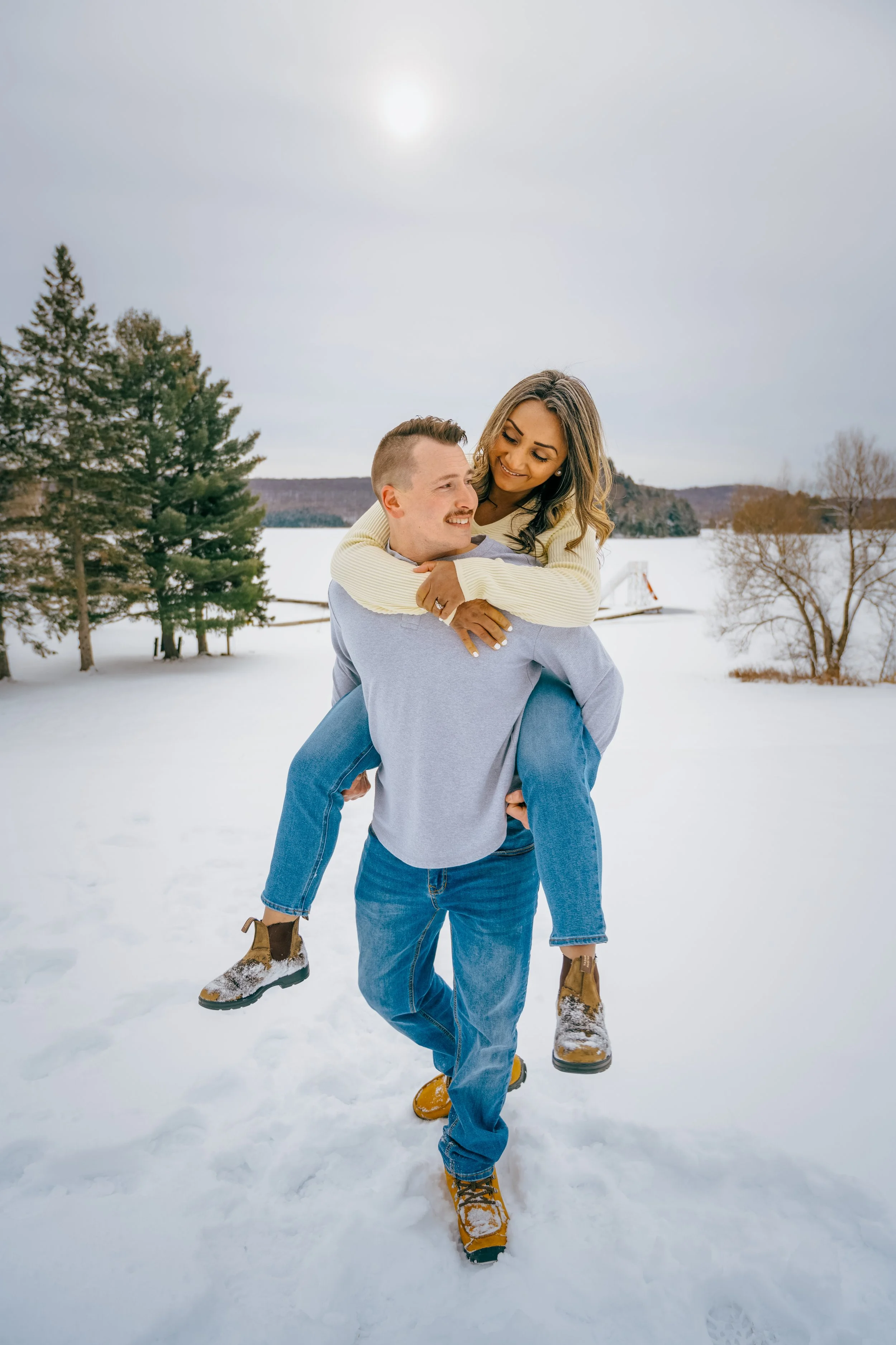 A couple outdoors in winter, with the woman on the man's back, smiling and looking at each other. Snow covers the ground, and there are trees and a lake in the background.