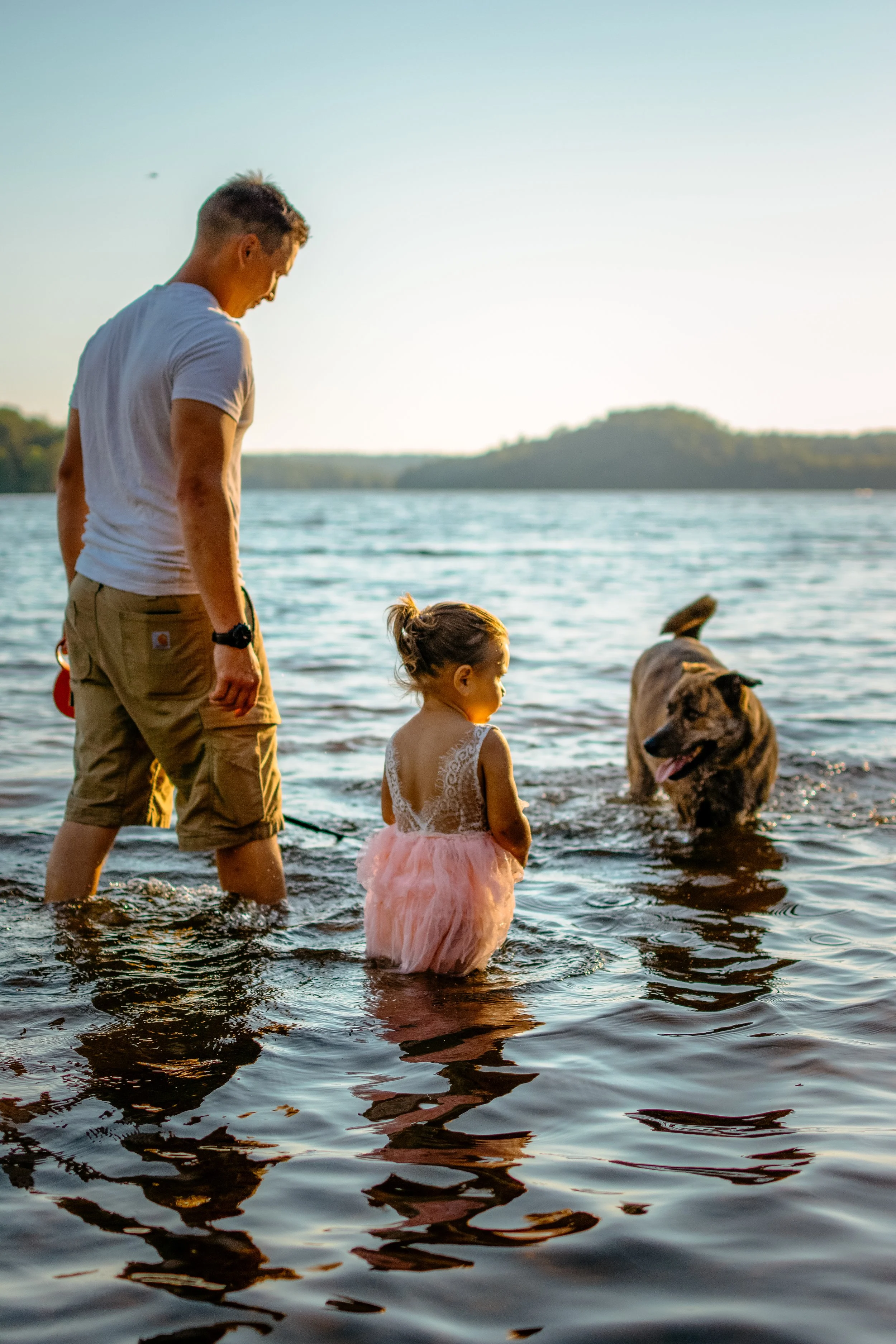 A man, a young girl, and a large dog wading in a river during sunset.
