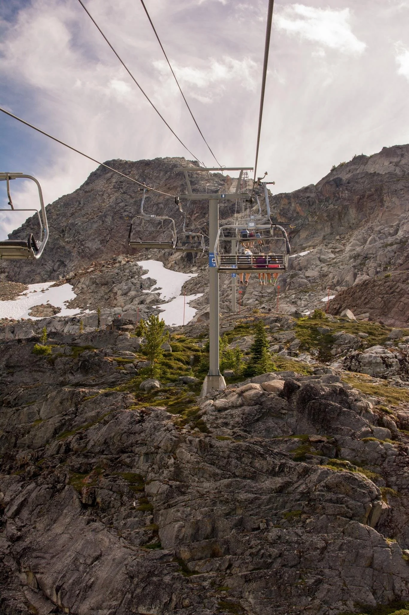 View of a ski lift with empty chairs on a rocky mountain with patches of snow and small green trees, under a cloudy sky.