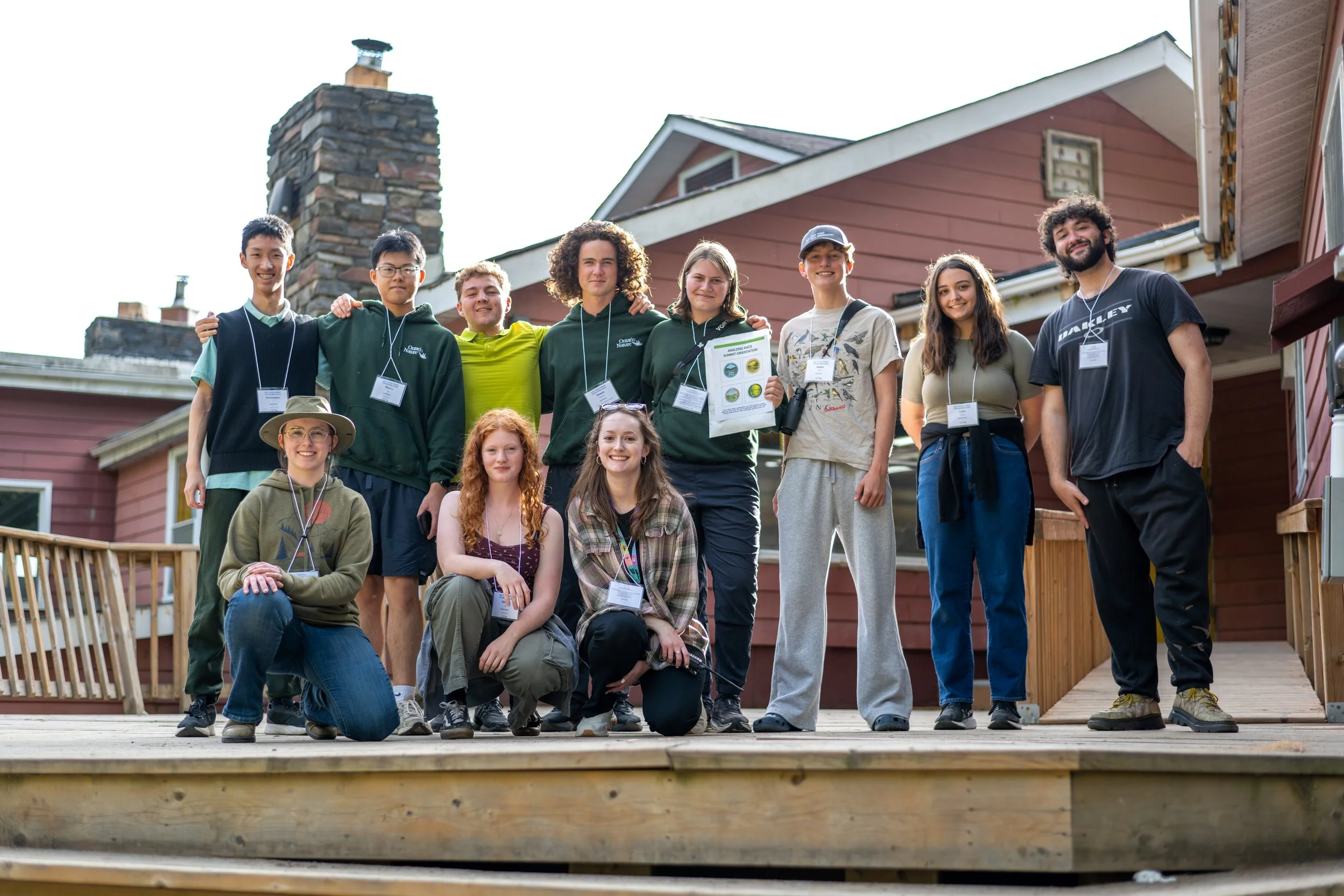 A group of young people, some with lanyards, posing on a wooden deck outside a house with pink siding, during daytime.