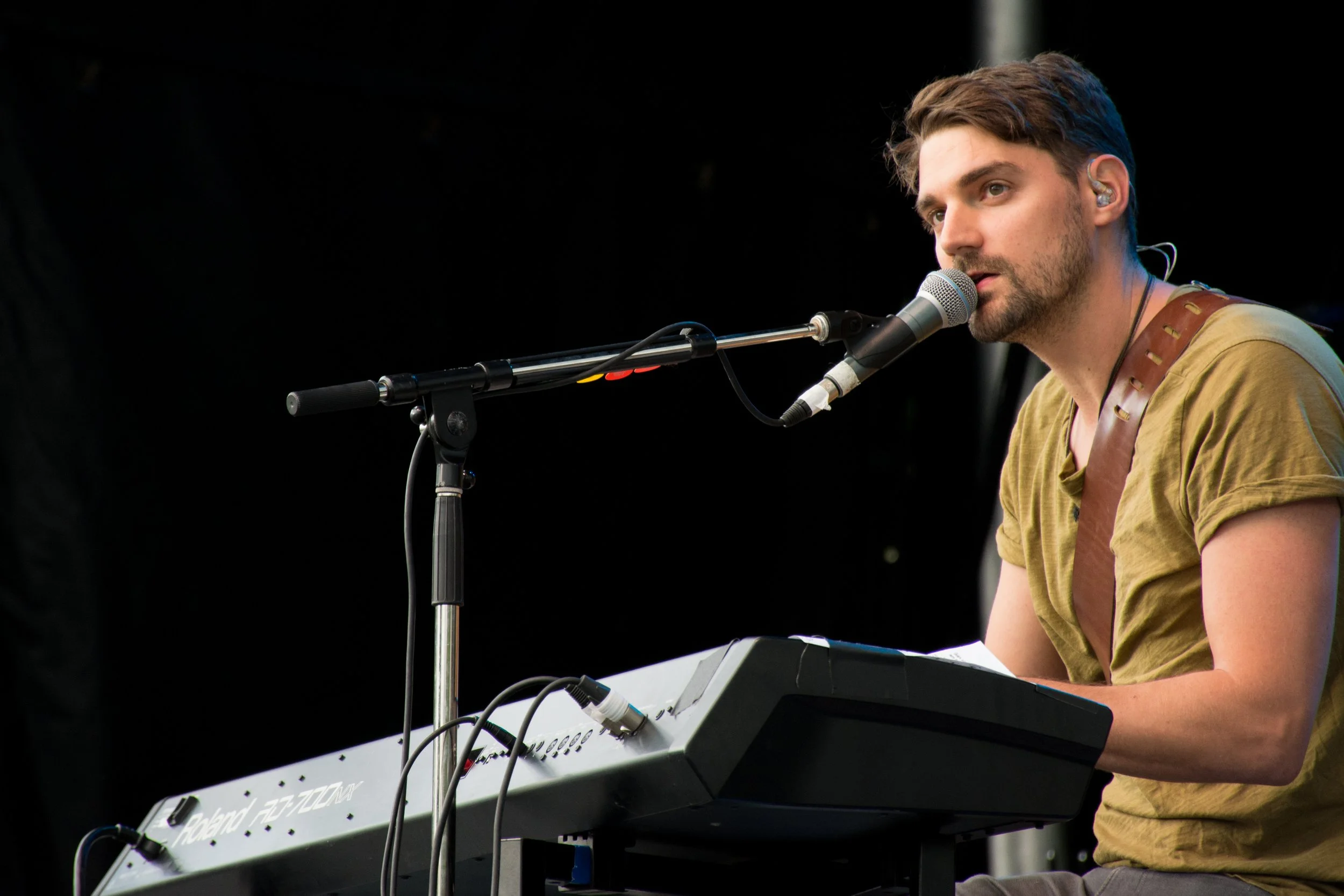 A man with brown hair and a beard plays an electronic keyboard and sings into a microphone on a dark stage.
